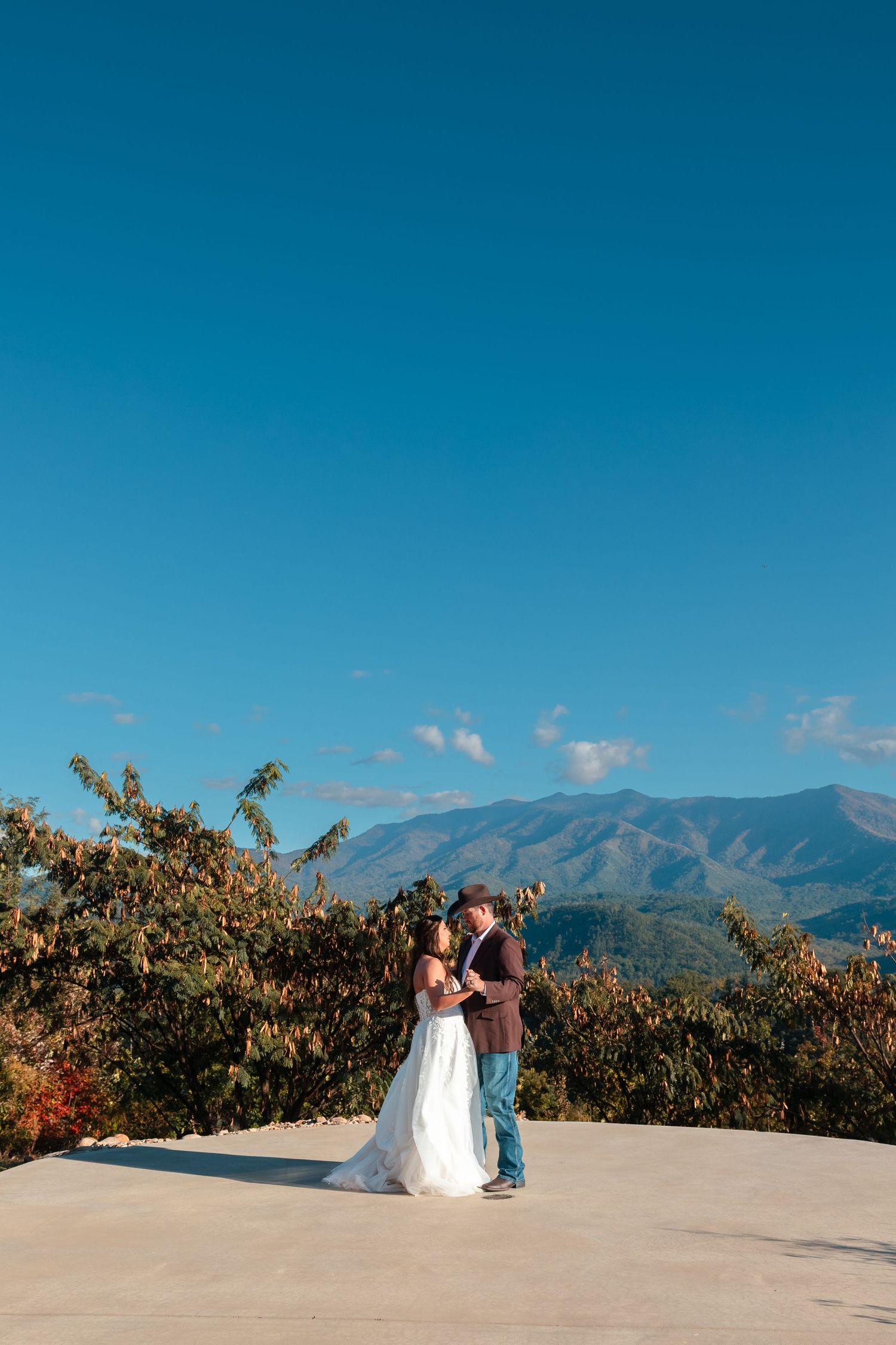 A couple stands on a scenic mountain overlook with blue skies and distant peaks in the background.