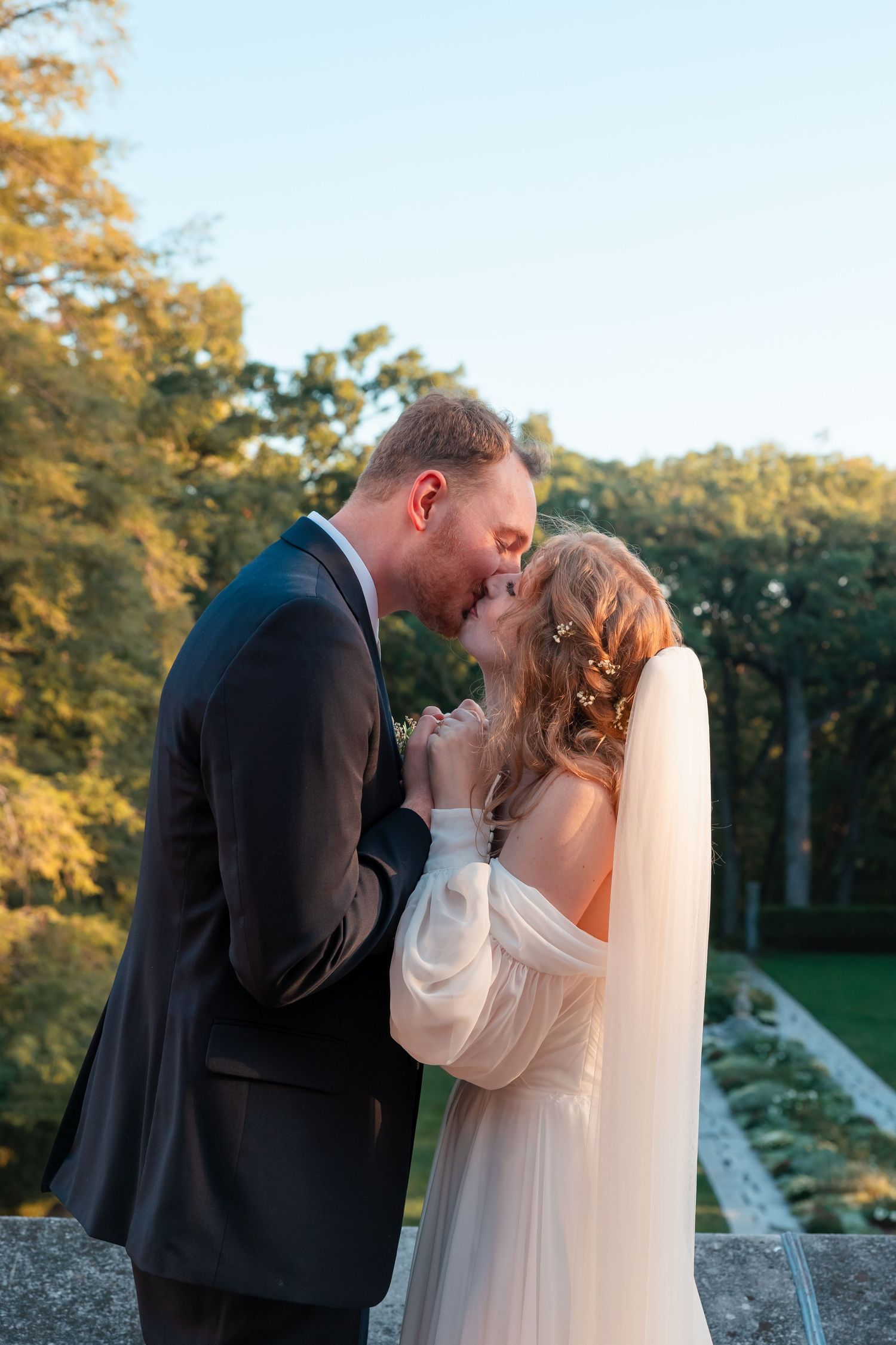A romantic wedding couple shares a tender kiss against a backdrop of autumn trees at sunset.