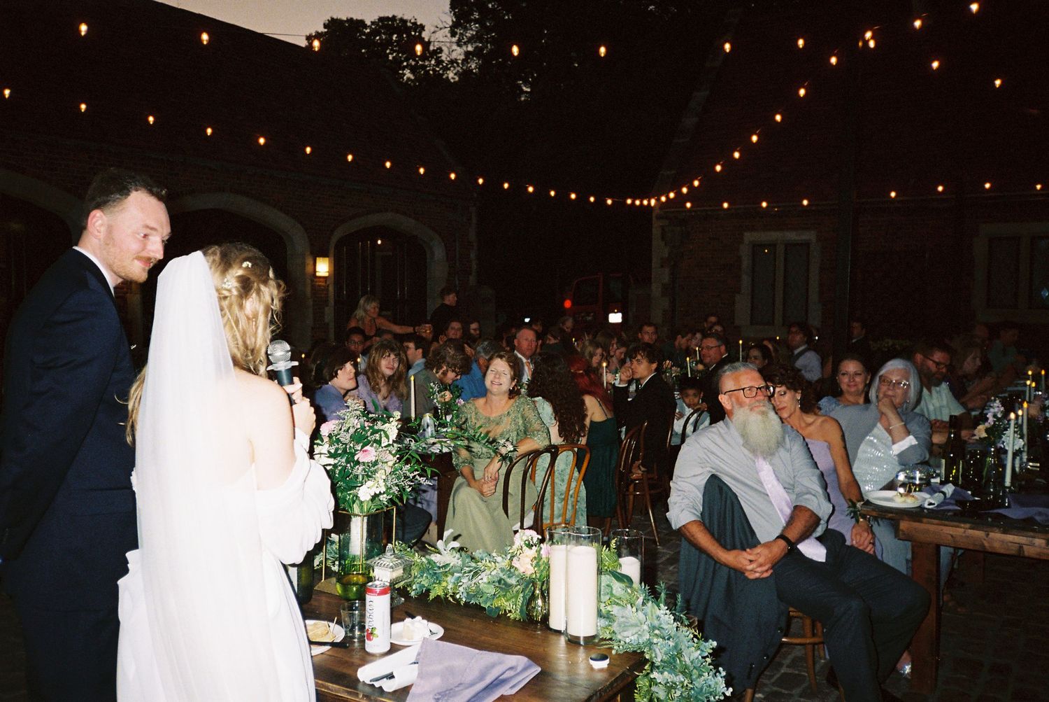 Wedding guests enjoy an evening reception under twinkling string lights in a rustic barn setting.