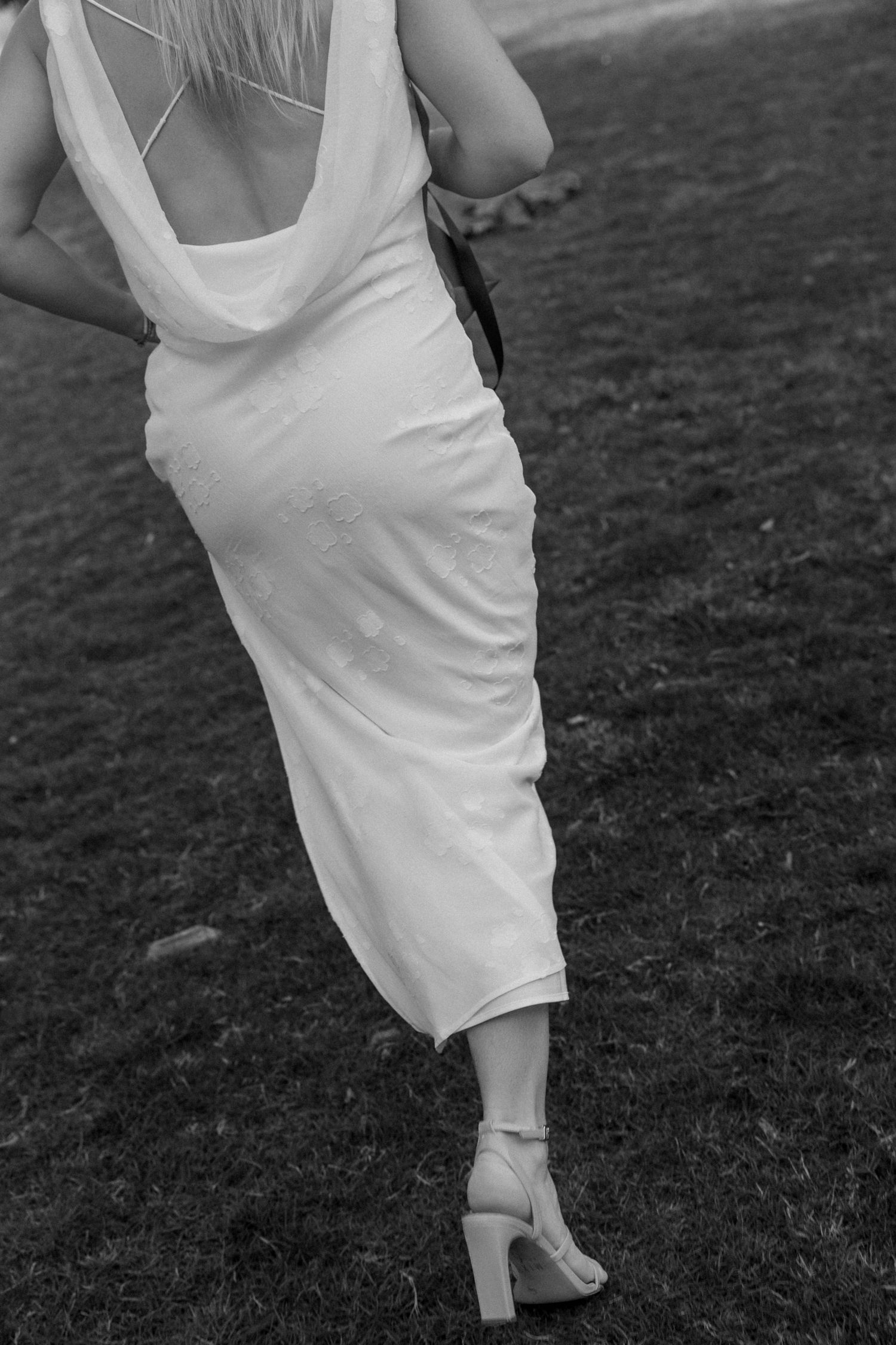 Black and white photo of a woman in a white backless dress and heels walking on grass.