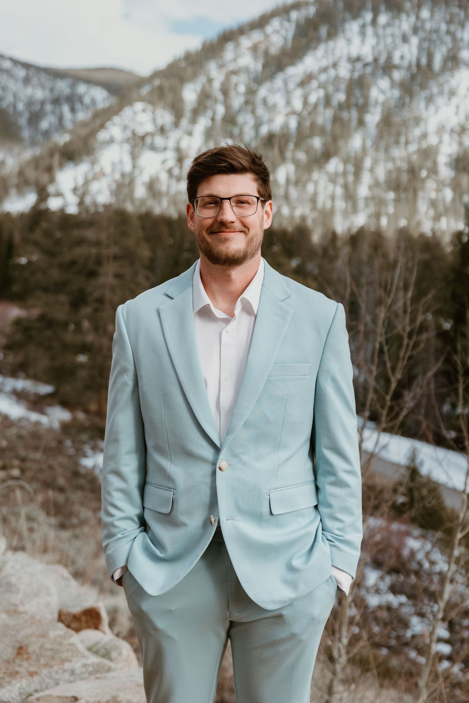 Person in a light blue suit jacket and glasses stands against a snowy mountain backdrop.