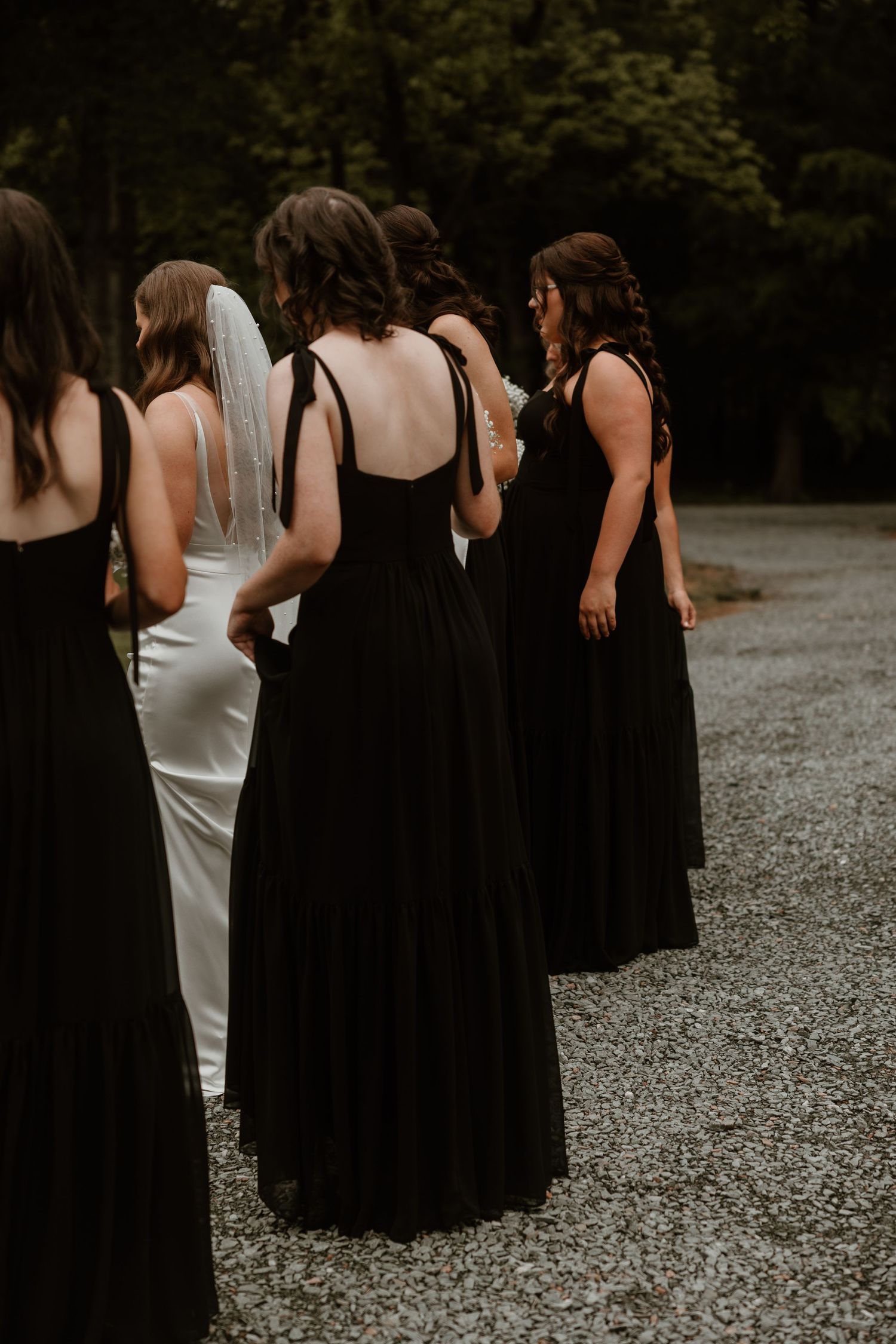 Bridesmaids in elegant black dresses walking together on a gravel path.