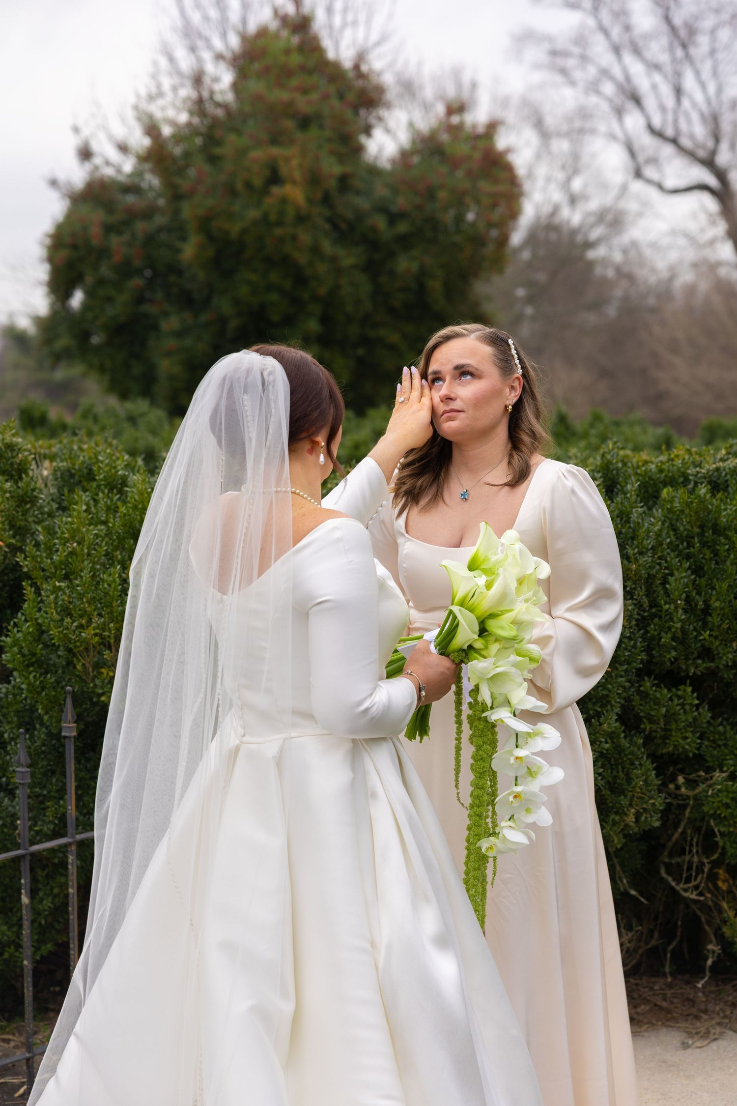 A group in white dresses and veils pose together outdoors with green bouquets against a hedge backdrop.