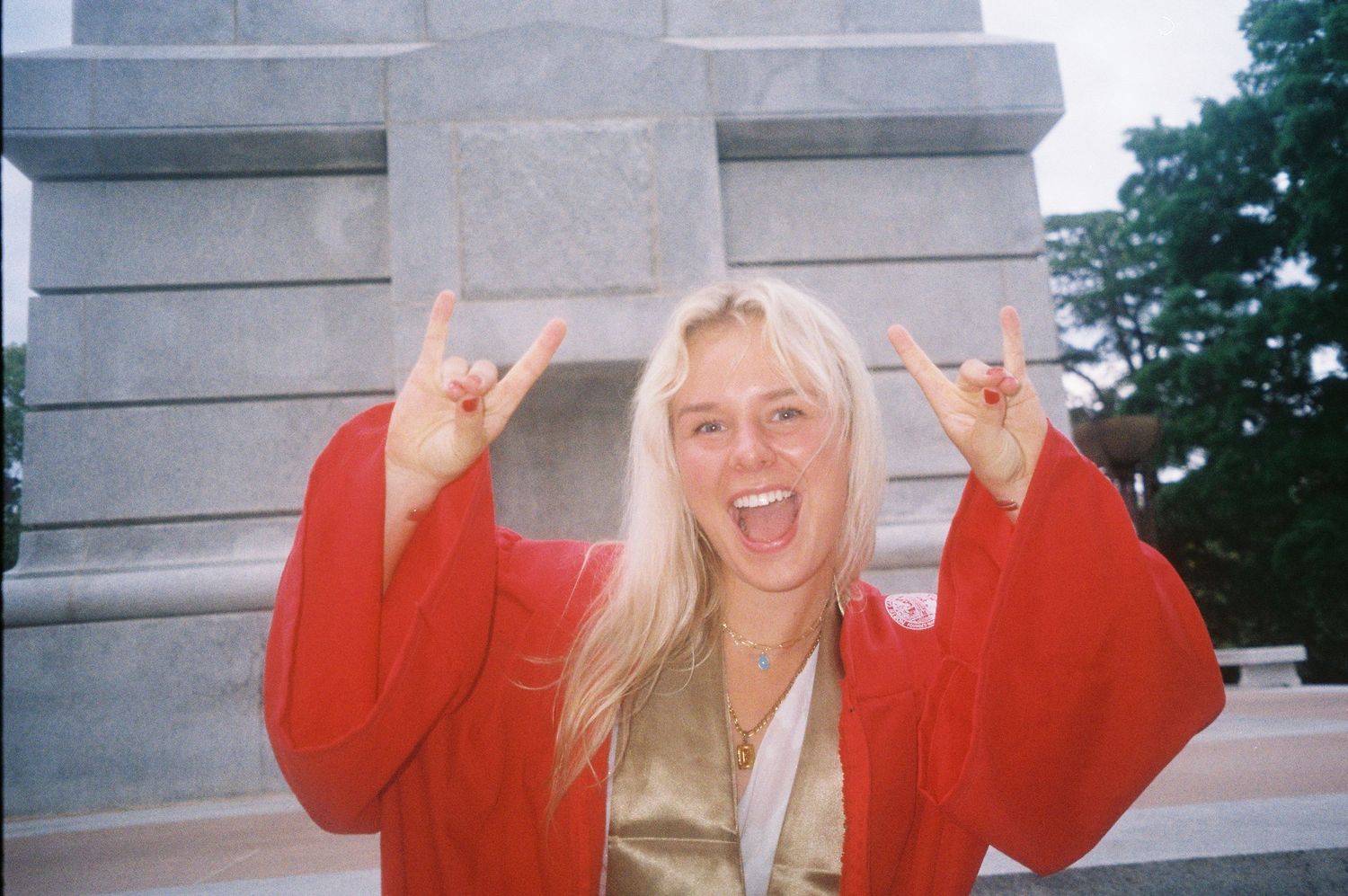 Smiling blonde woman in red graduation robe making rock on hand gestures in front of a stone monument.