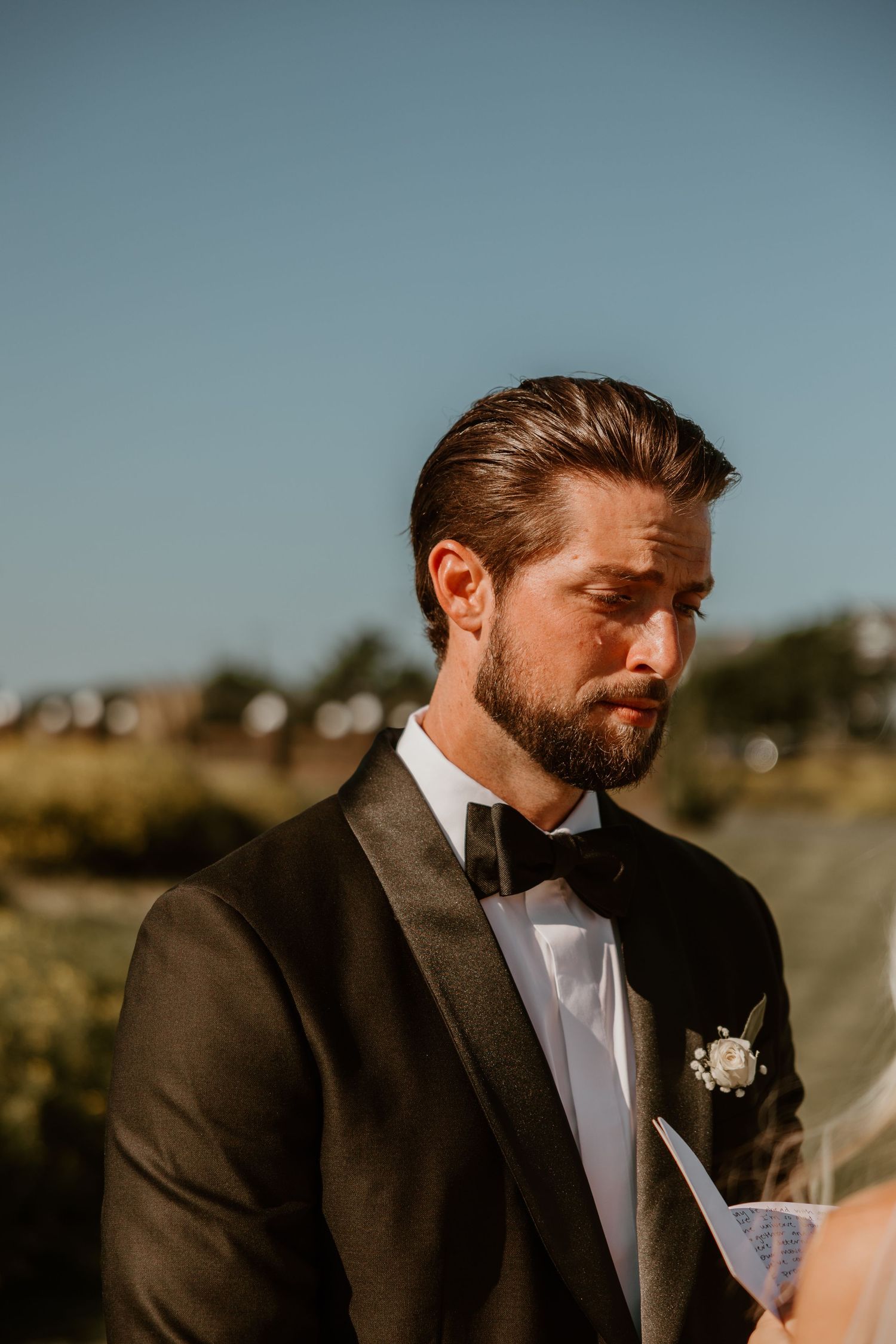 Groom in classic black tuxedo reads wedding vows in warm natural lighting.