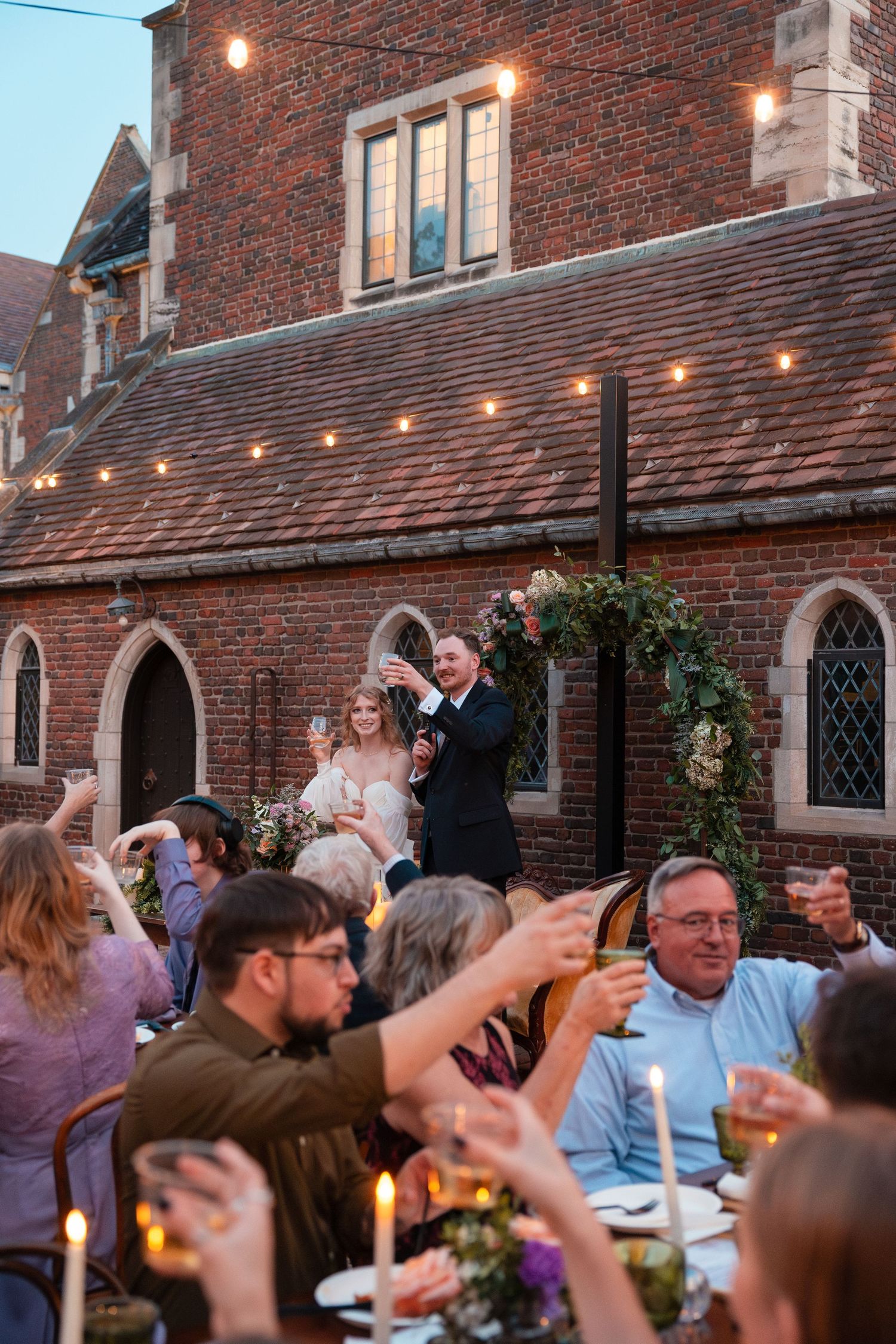 Wedding reception guests raising their glasses in a toast under string lights at a brick venue.