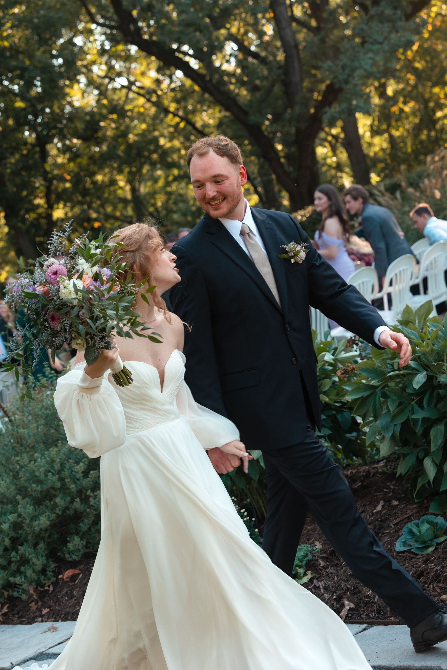 Bride and groom share playful dancing moments outdoors among garden greenery.