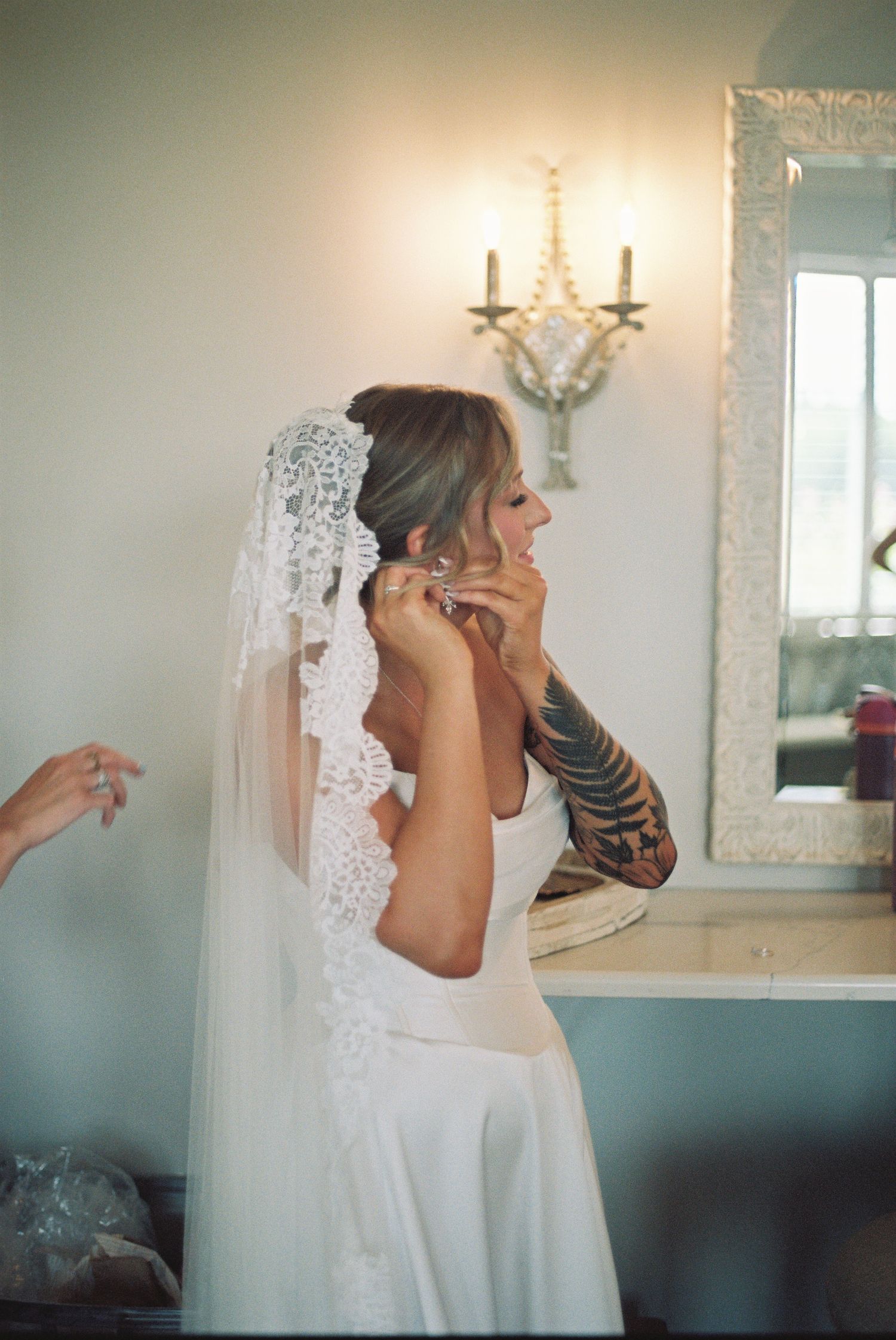A bride in a white lace veil and sleeveless dress with visible arm tattoos prepares on her wedding day.