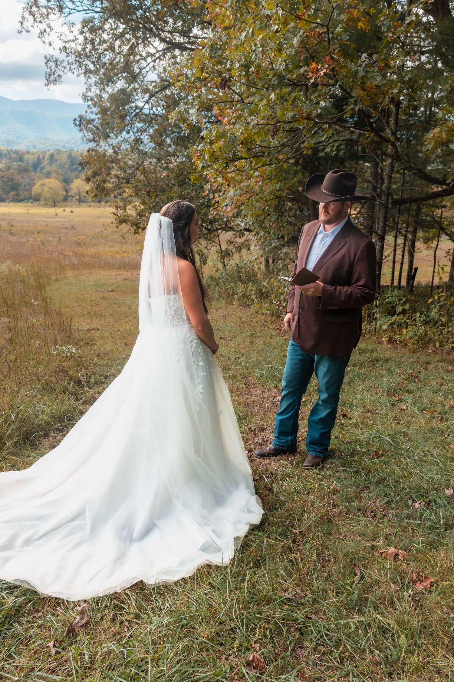 Wedding dress train spreads across autumn grass as couple poses in rural setting.