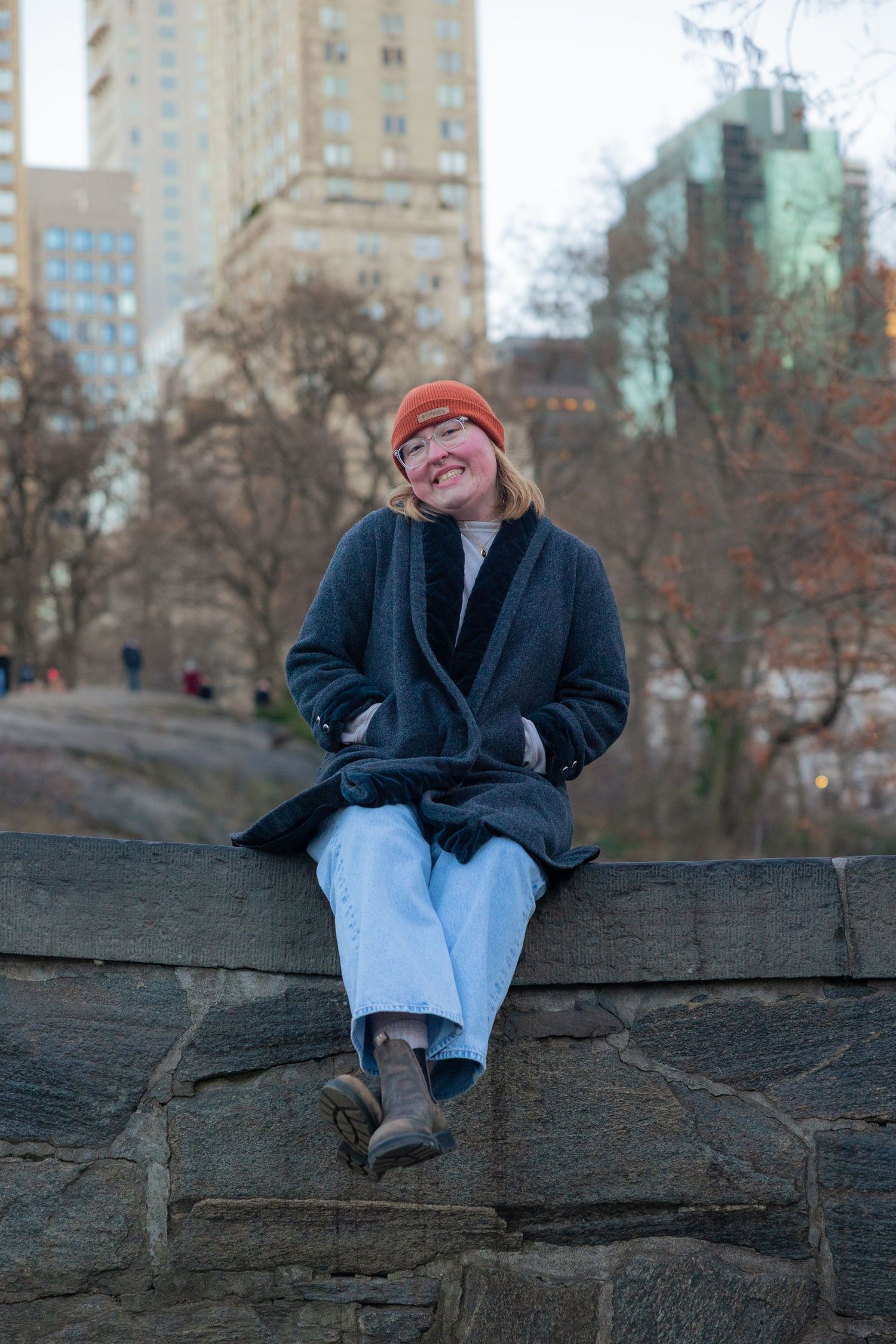 Person wearing a red beanie and winter coat sits casually on a large rock formation in an urban park setting.