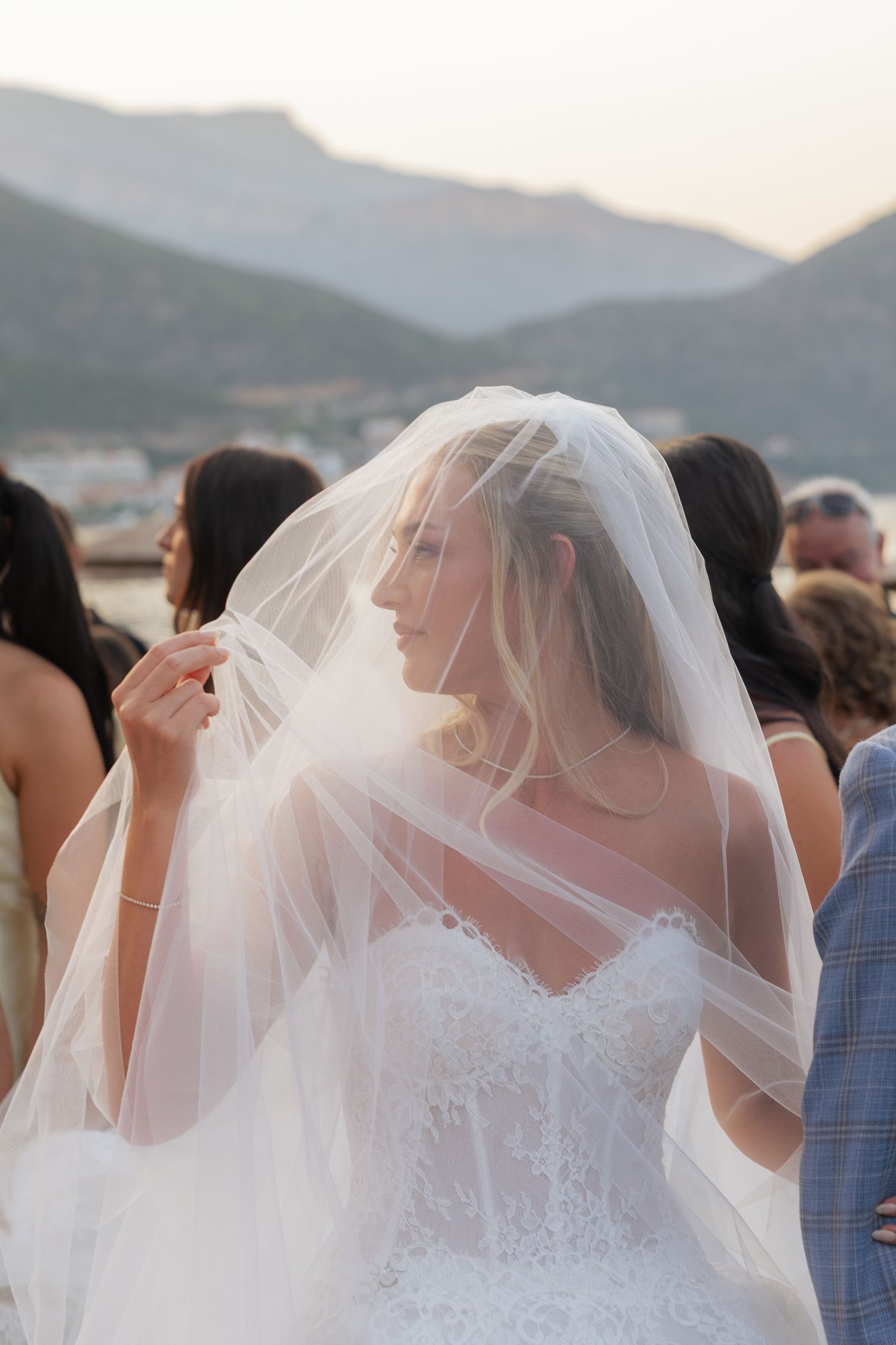 A bride in a white lace gown and veil poses against a scenic mountain backdrop during sunset.
