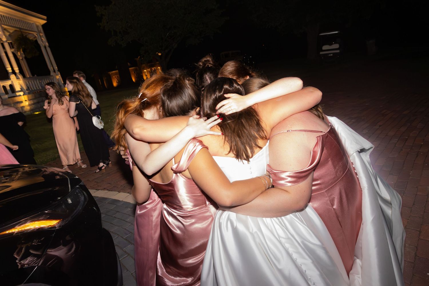 A group of wedding guests embrace in a circle during an evening celebration.