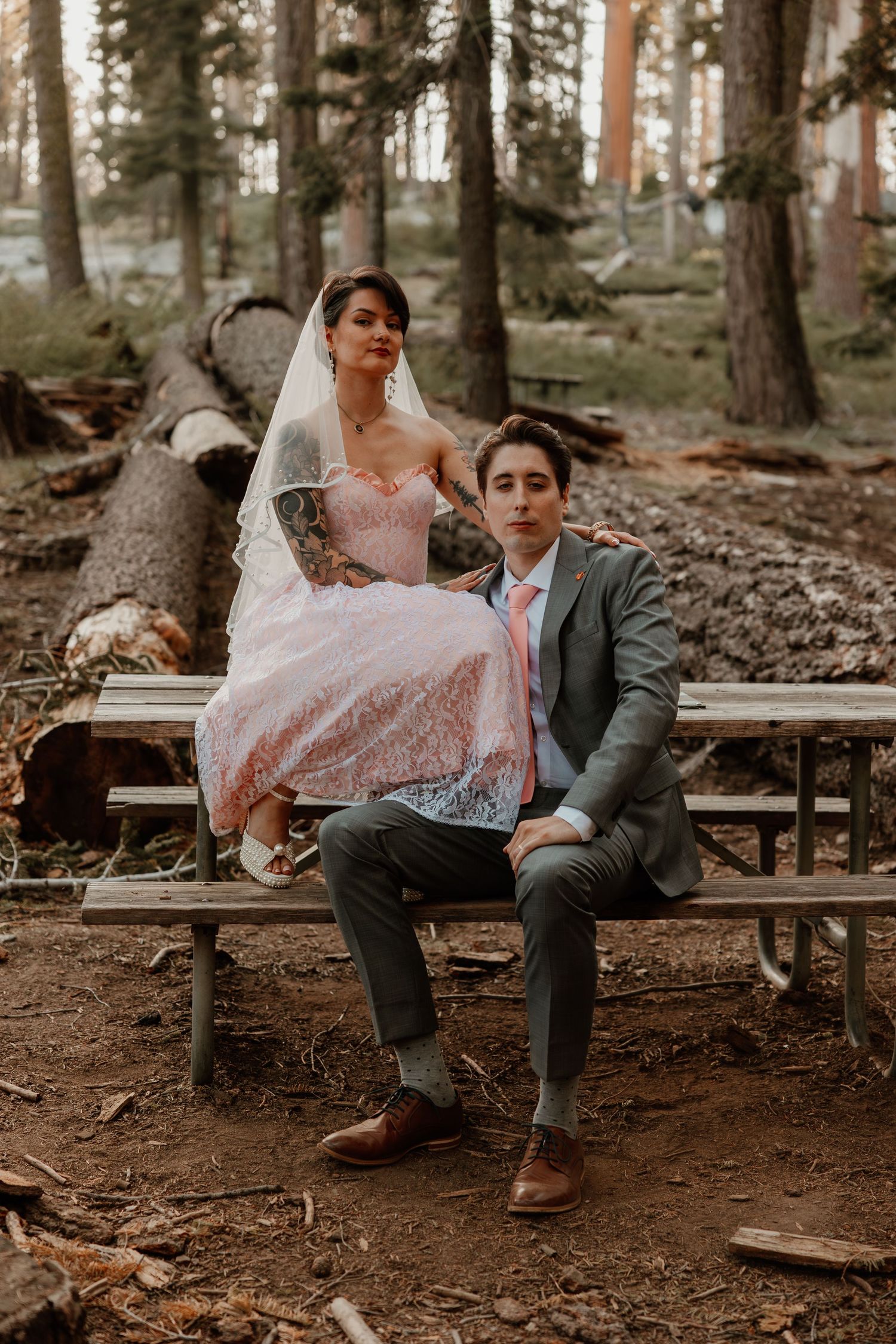 Wedding couple poses on rustic wooden bench in forest setting.