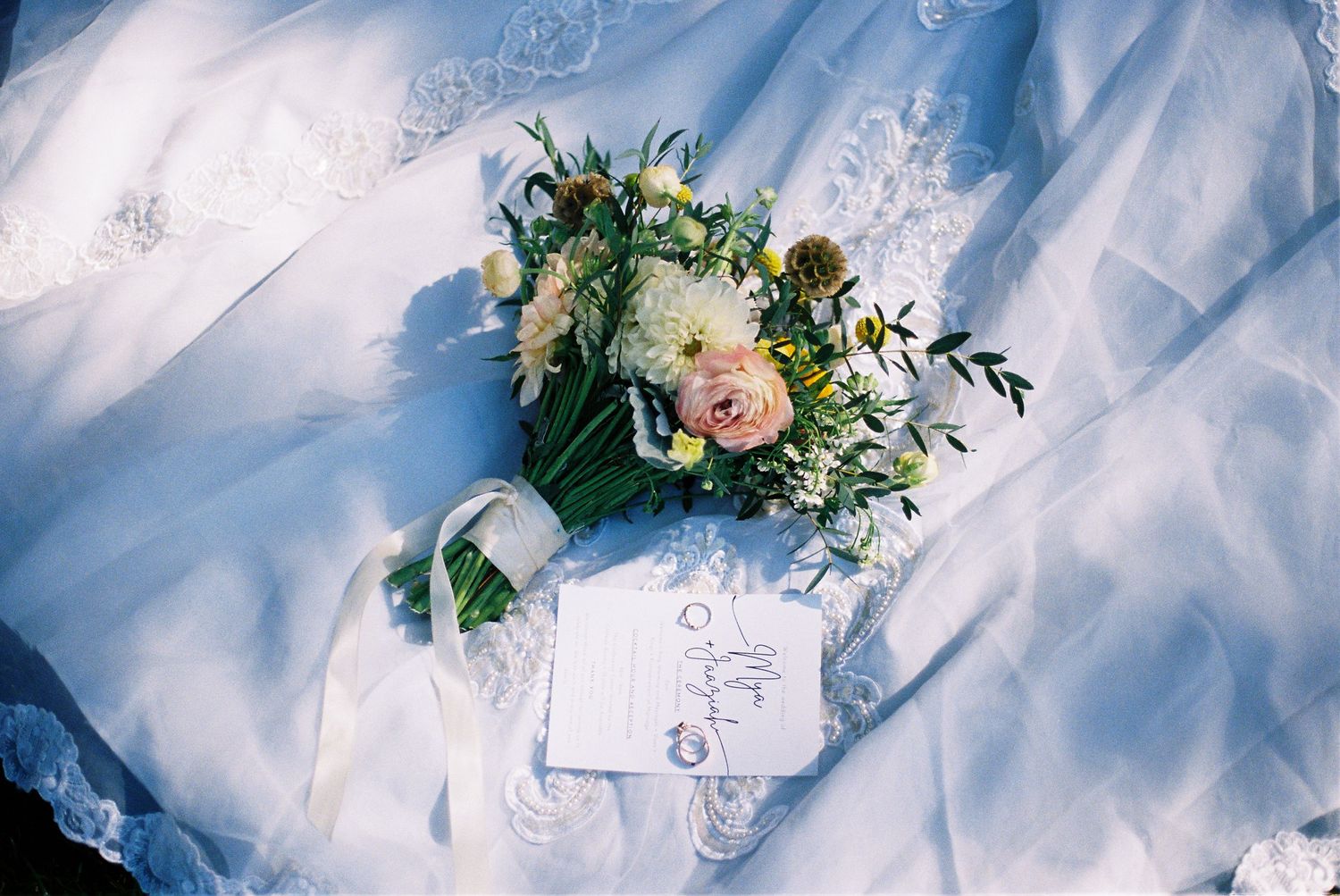 A wedding bouquet with peach roses and greenery laying on white lace fabric with a note card.