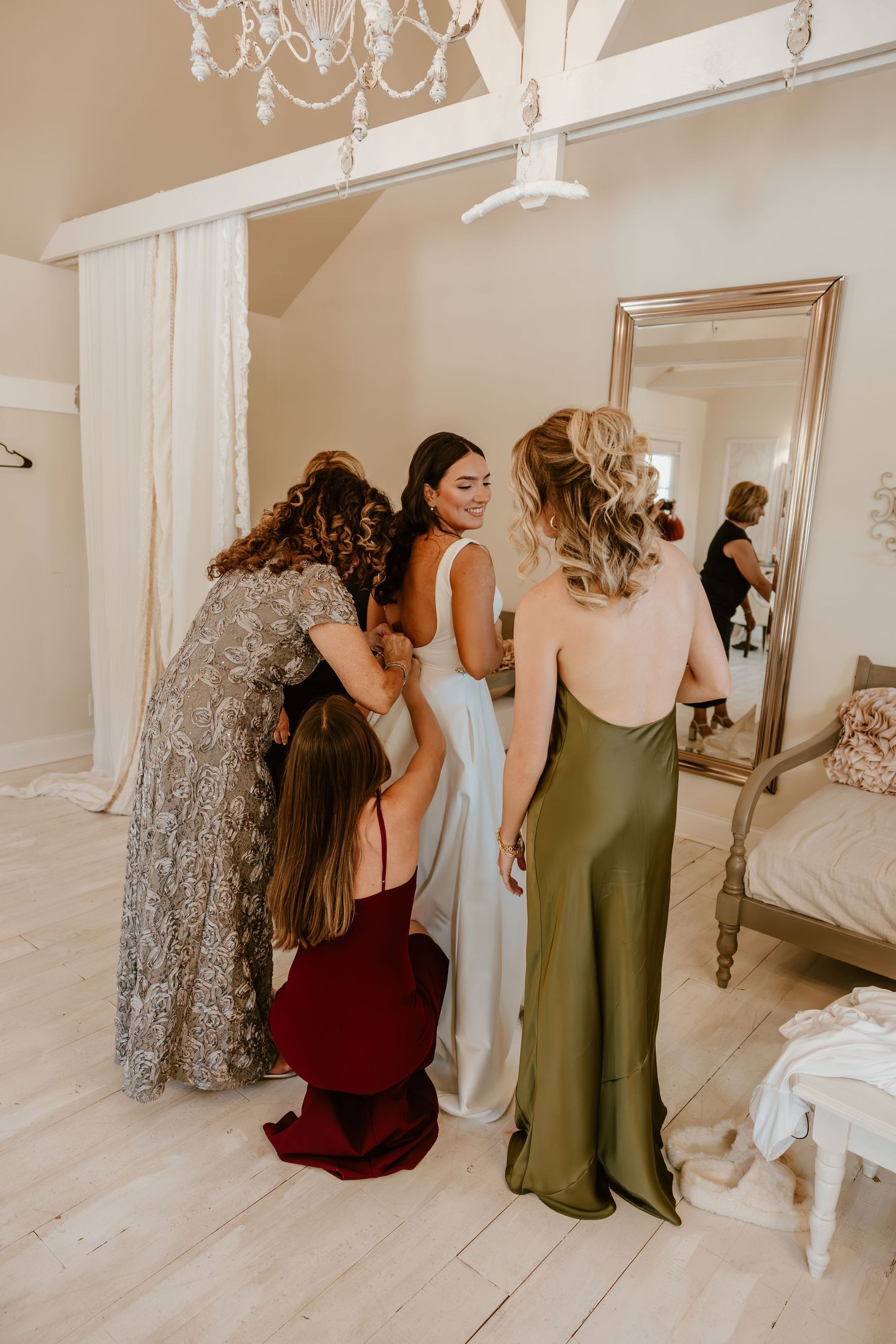 Wedding party in various colored dresses preparing in an elegant white room with chandelier.