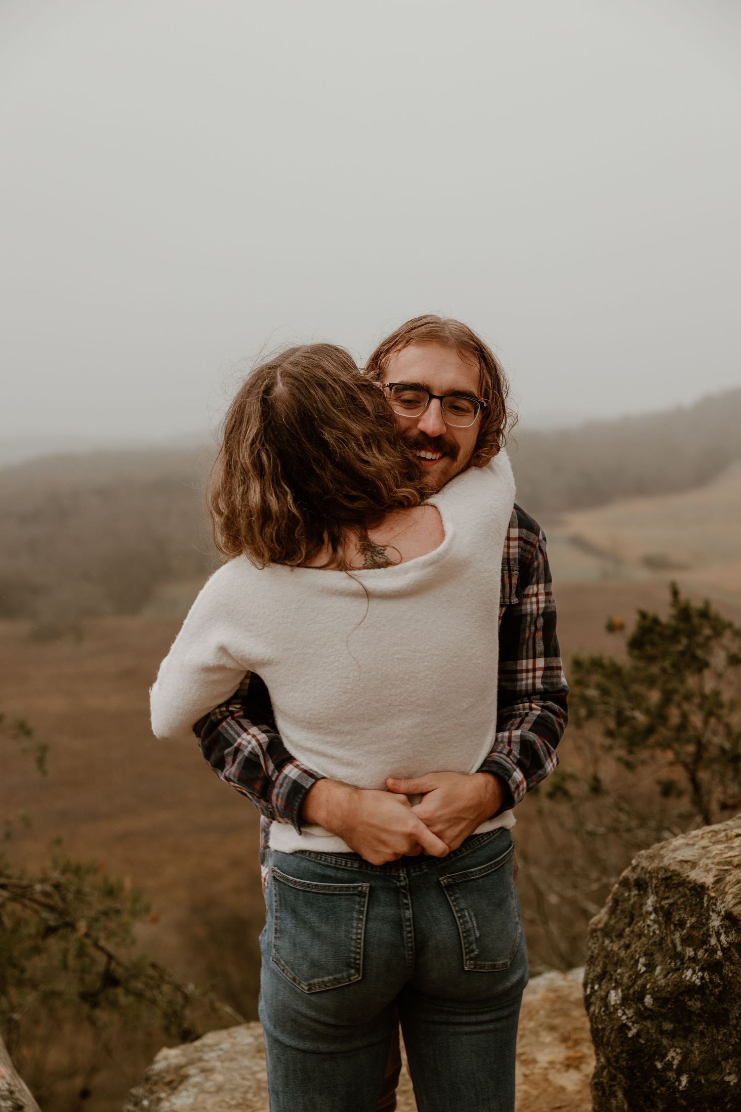 Intimate embrace captured against misty mountain backdrop on rocky overlook.