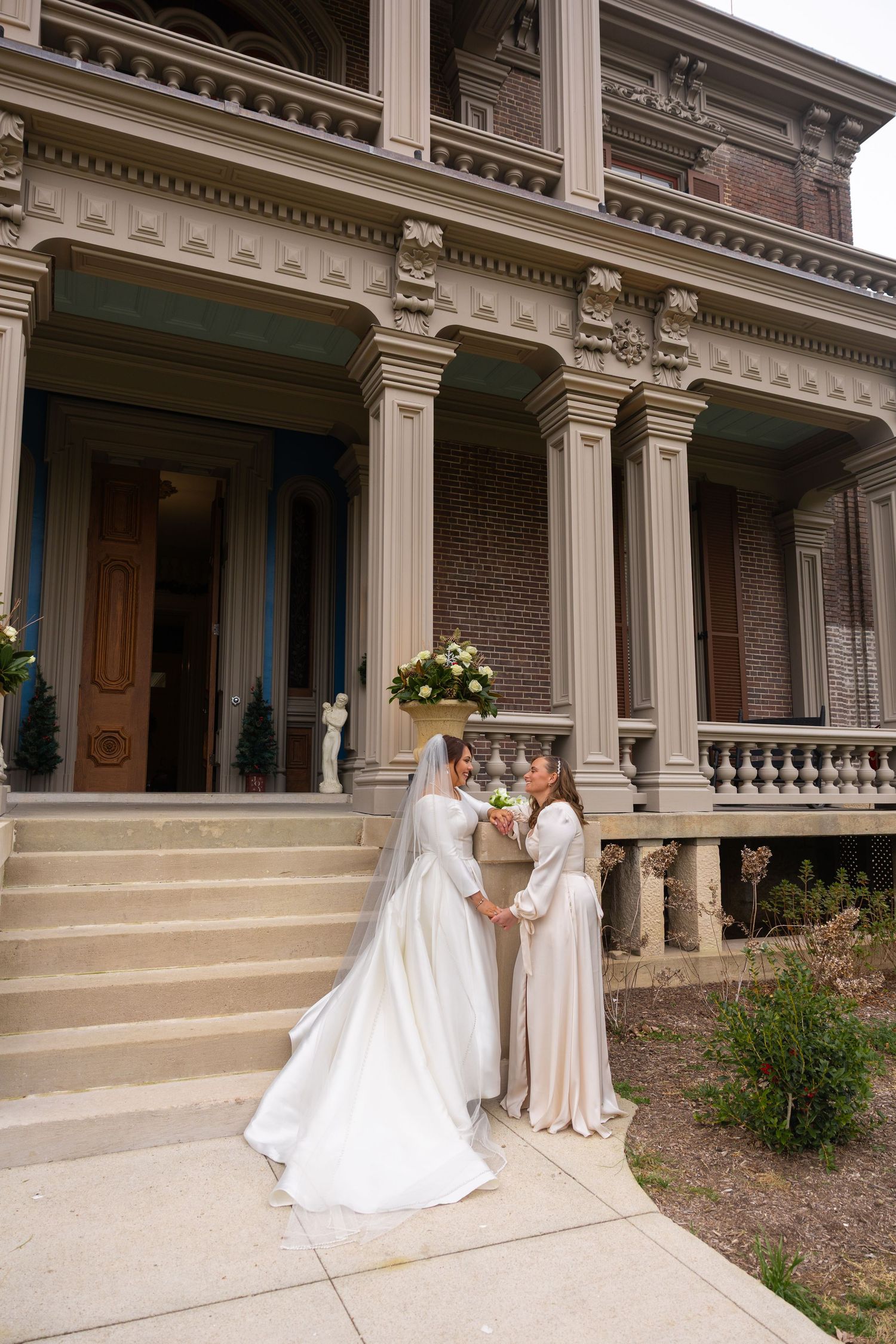 Two people in elegant white gowns pose together on the steps of a historic building with classical columns.