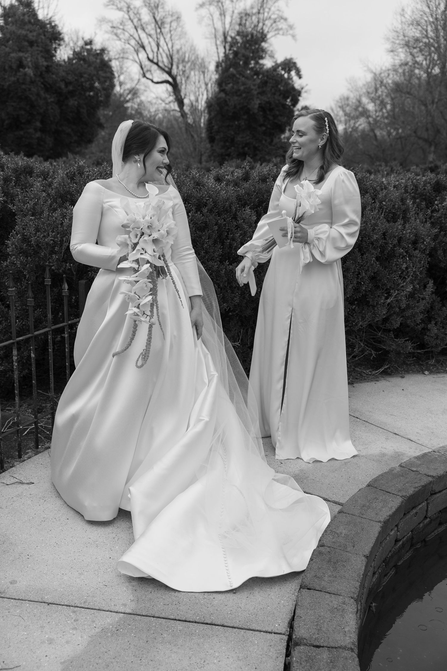 Two people in elegant white dresses stand together on stone steps in a black and white outdoor photo series.