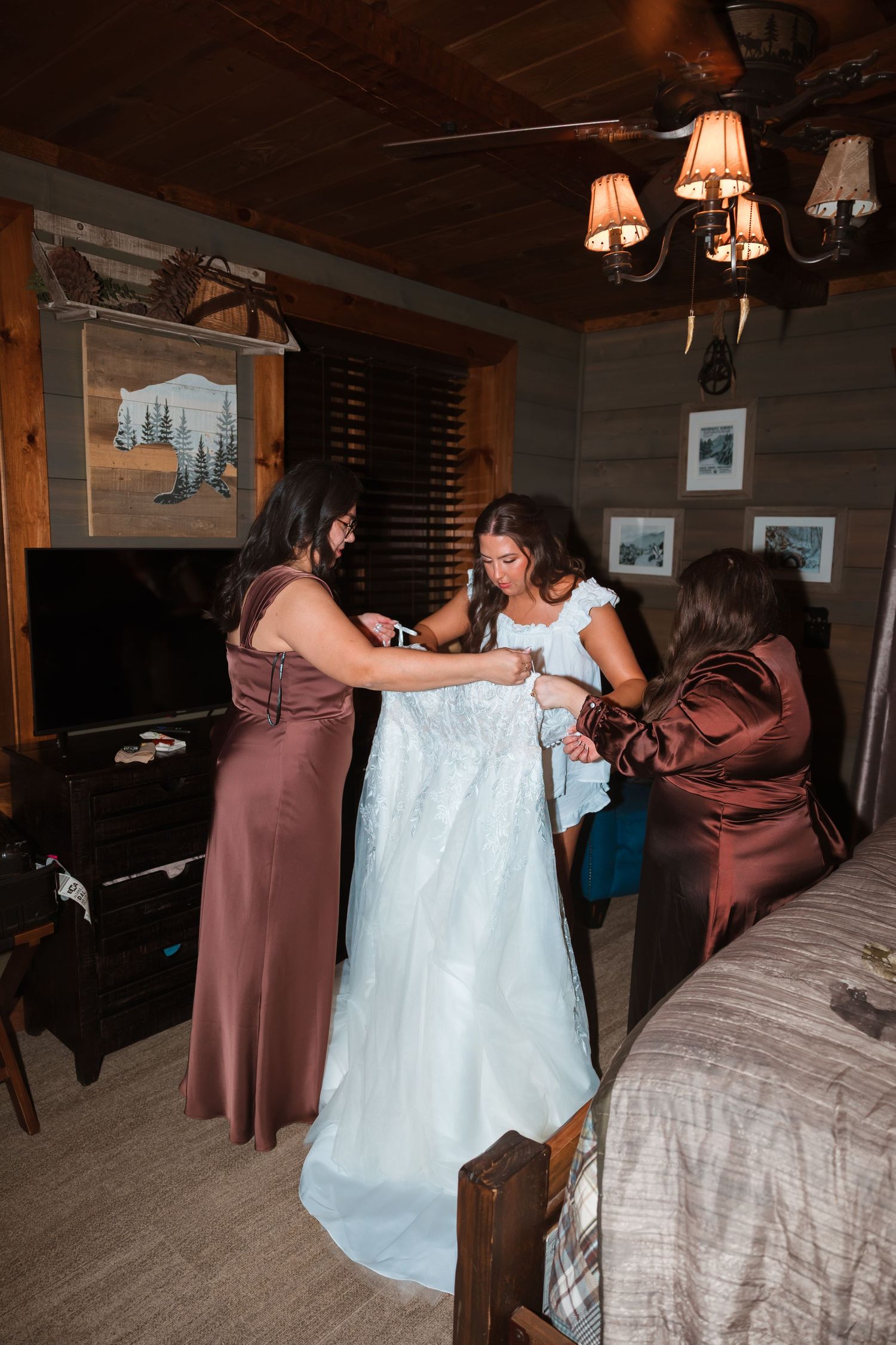Wedding dress being fitted and adjusted in a dimly lit rustic cabin setting.