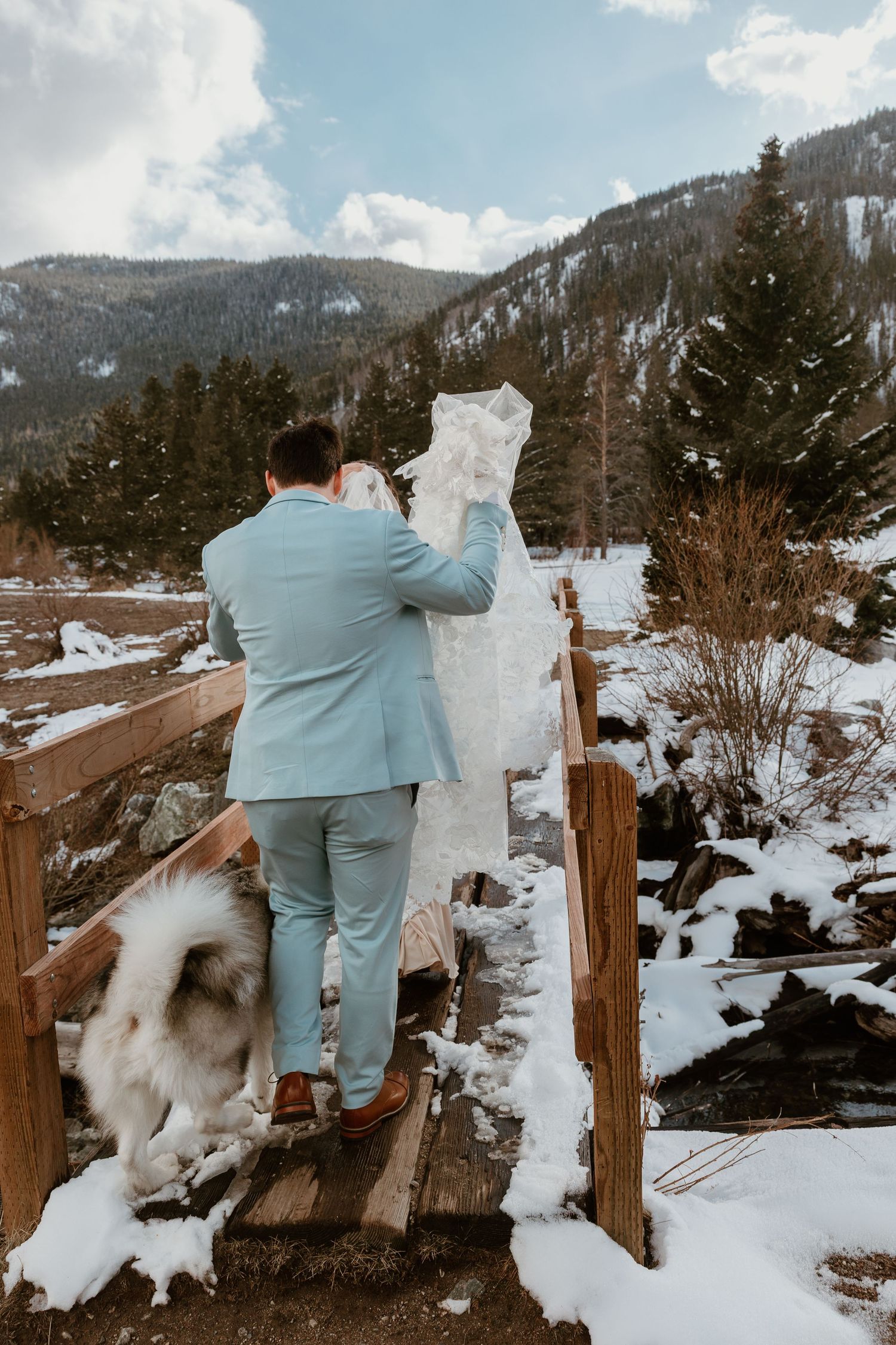 A couple in wedding attire dances and twirls on a snow-covered mountain path.