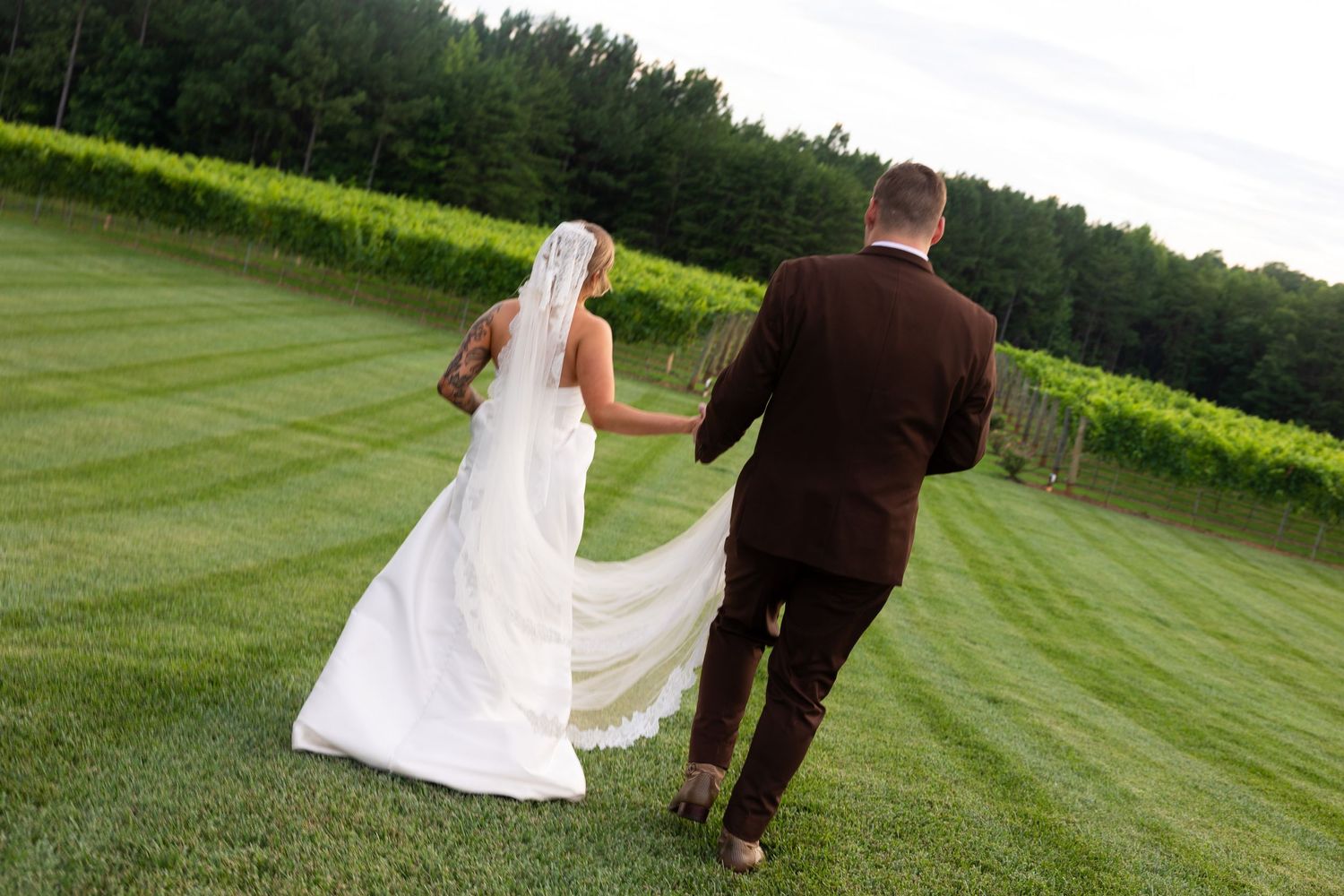A couple in wedding attire walks together on a lush green lawn during their outdoor celebration.