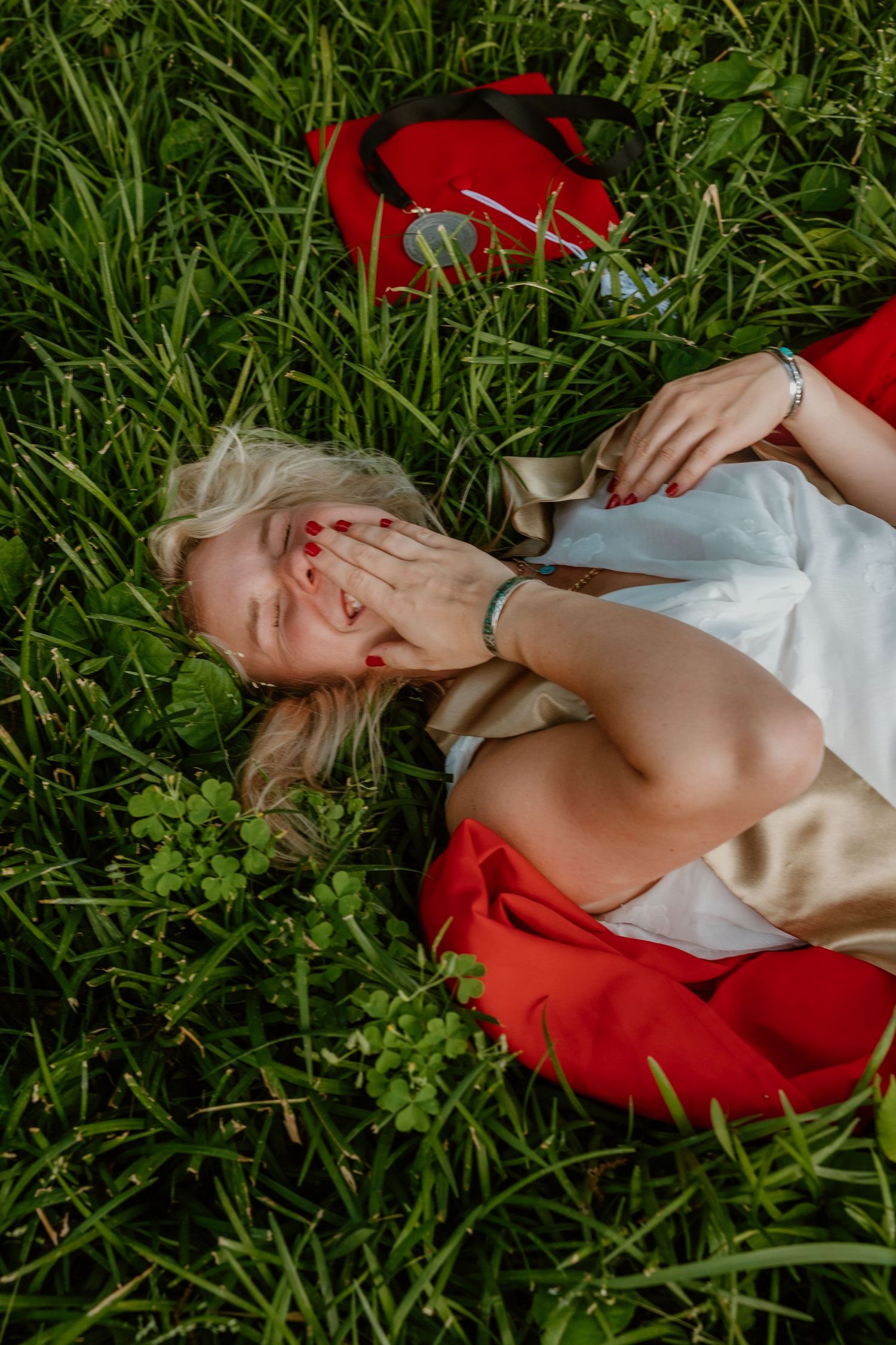 Blonde woman lying on green grass laughing, wearing white dress with red pillow and bracelet.