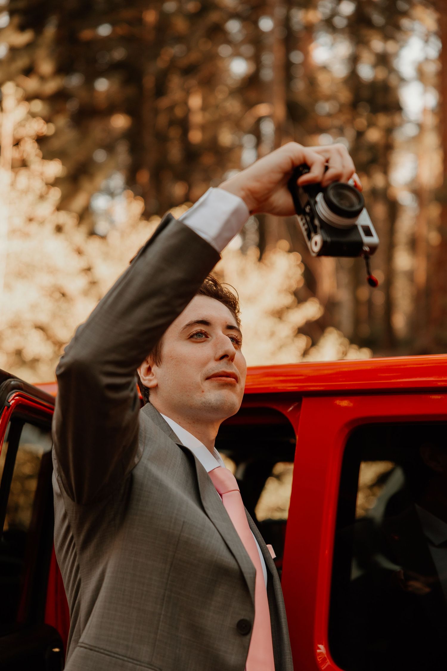 Person in grey suit holding up a vintage camera near a red vehicle in the forest.