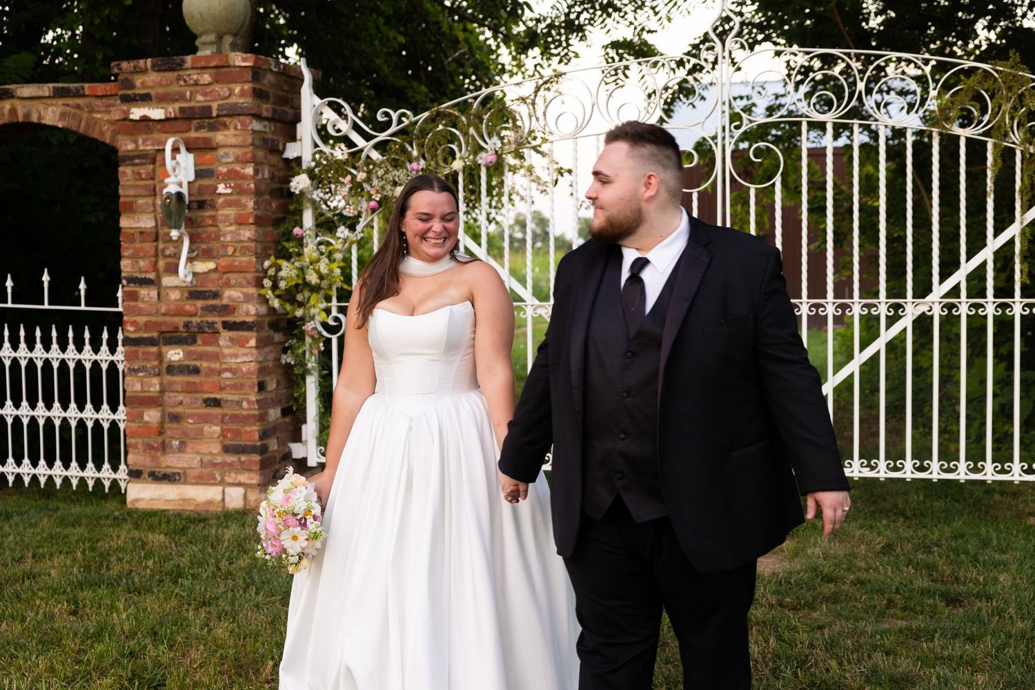 A couple in formal wedding attire walks together on a sunny day near a brick wall and white iron gate.