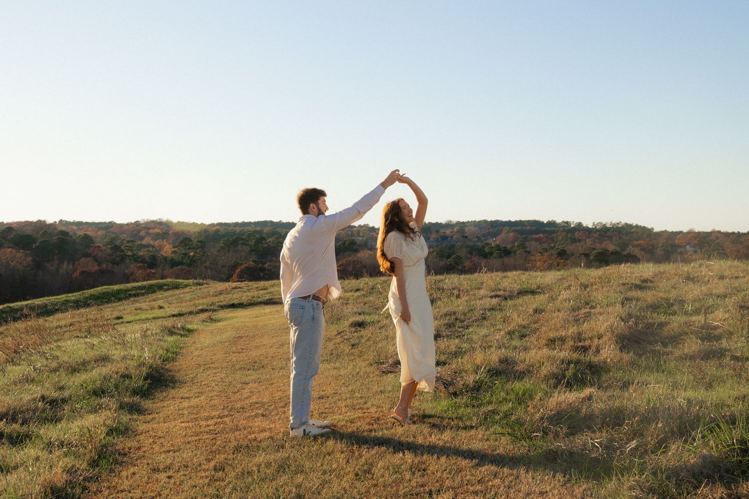 Couple in white outfits walking together through golden grass fields at sunset, holding hands and embracing.