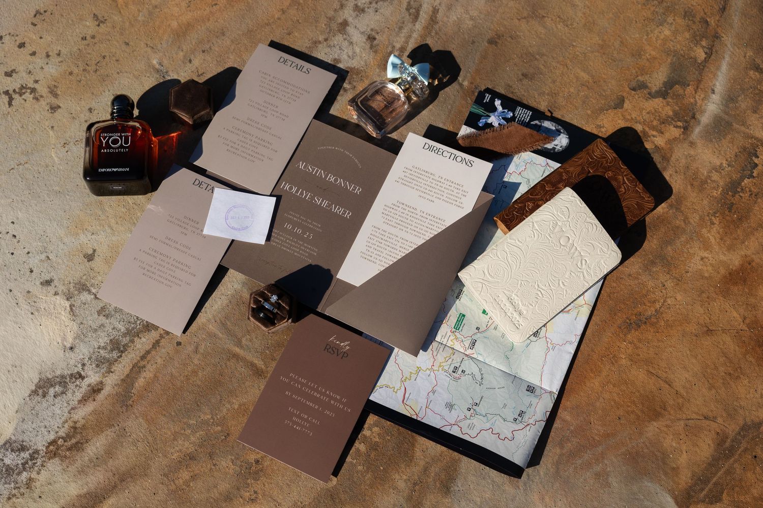 Wedding stationery set with brown invitations, envelopes and accessories laid out on a textured stone surface.