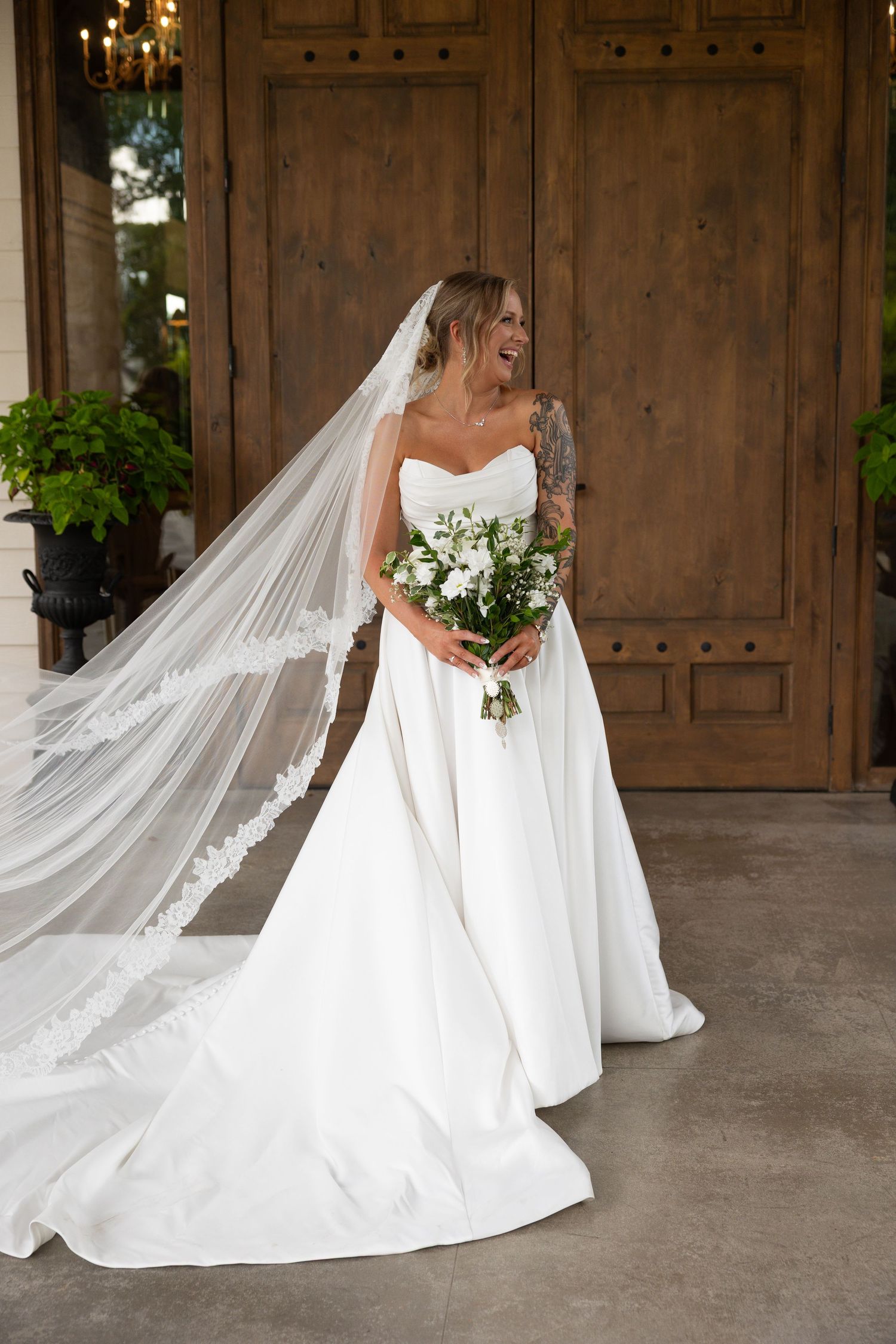A bride in a strapless white gown with long veil poses by wooden doors holding white floral bouquet.