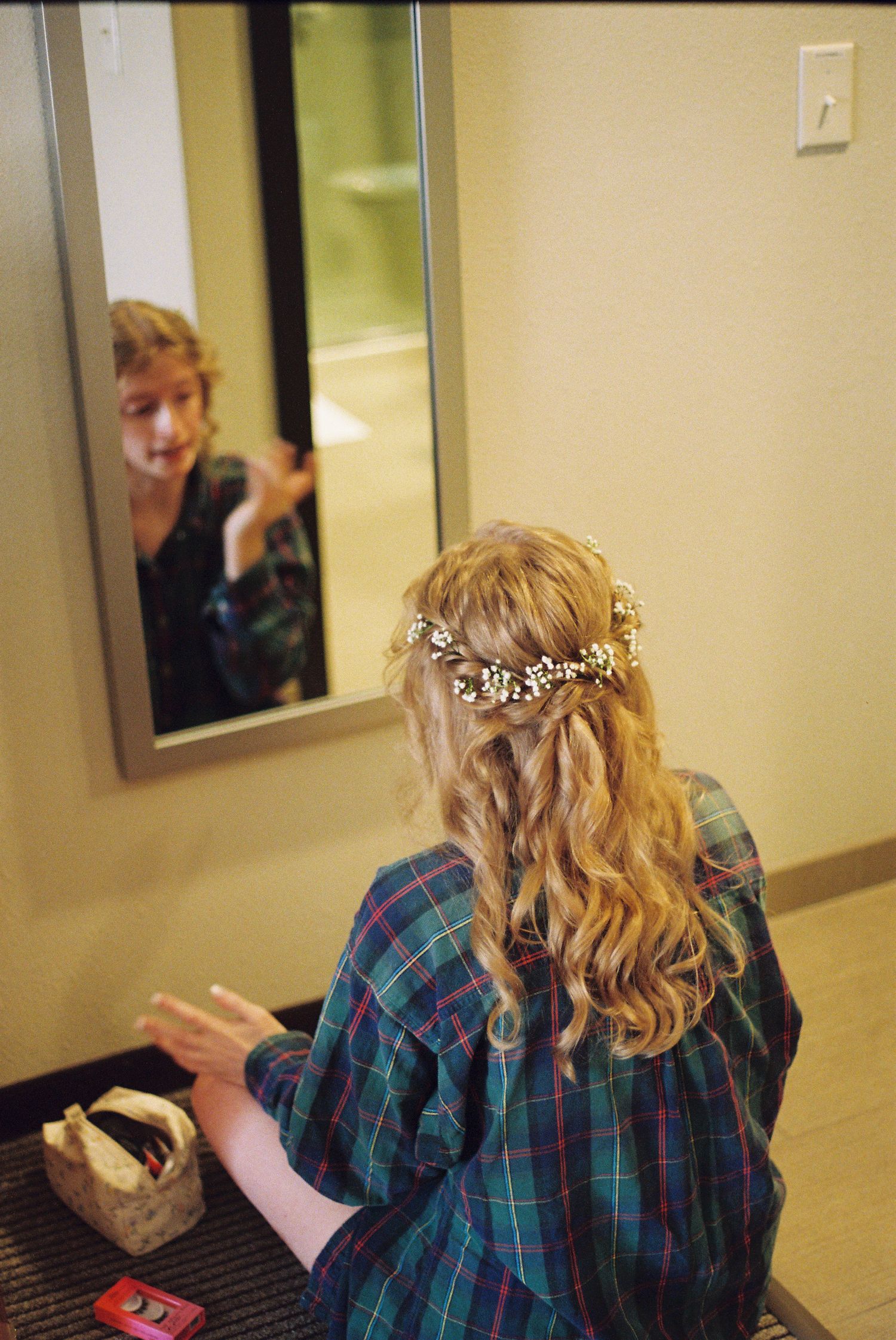 Bride getting ready in plaid robe with delicate floral hairpiece, seen from behind in mirror reflection.