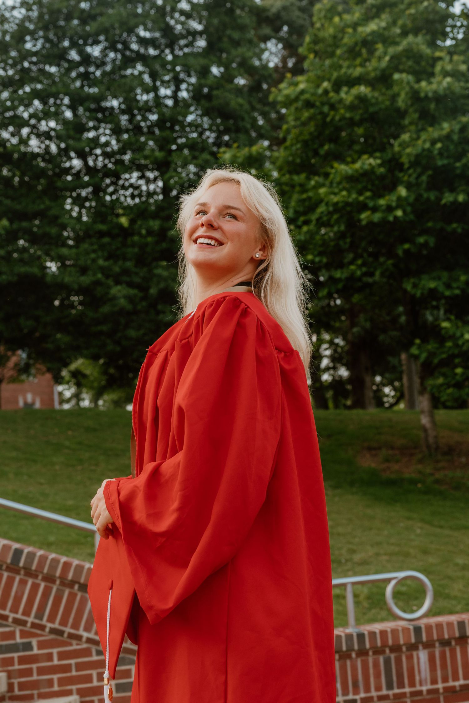 Smiling blonde woman in red graduation gown standing outdoors on campus with lush green trees in background.