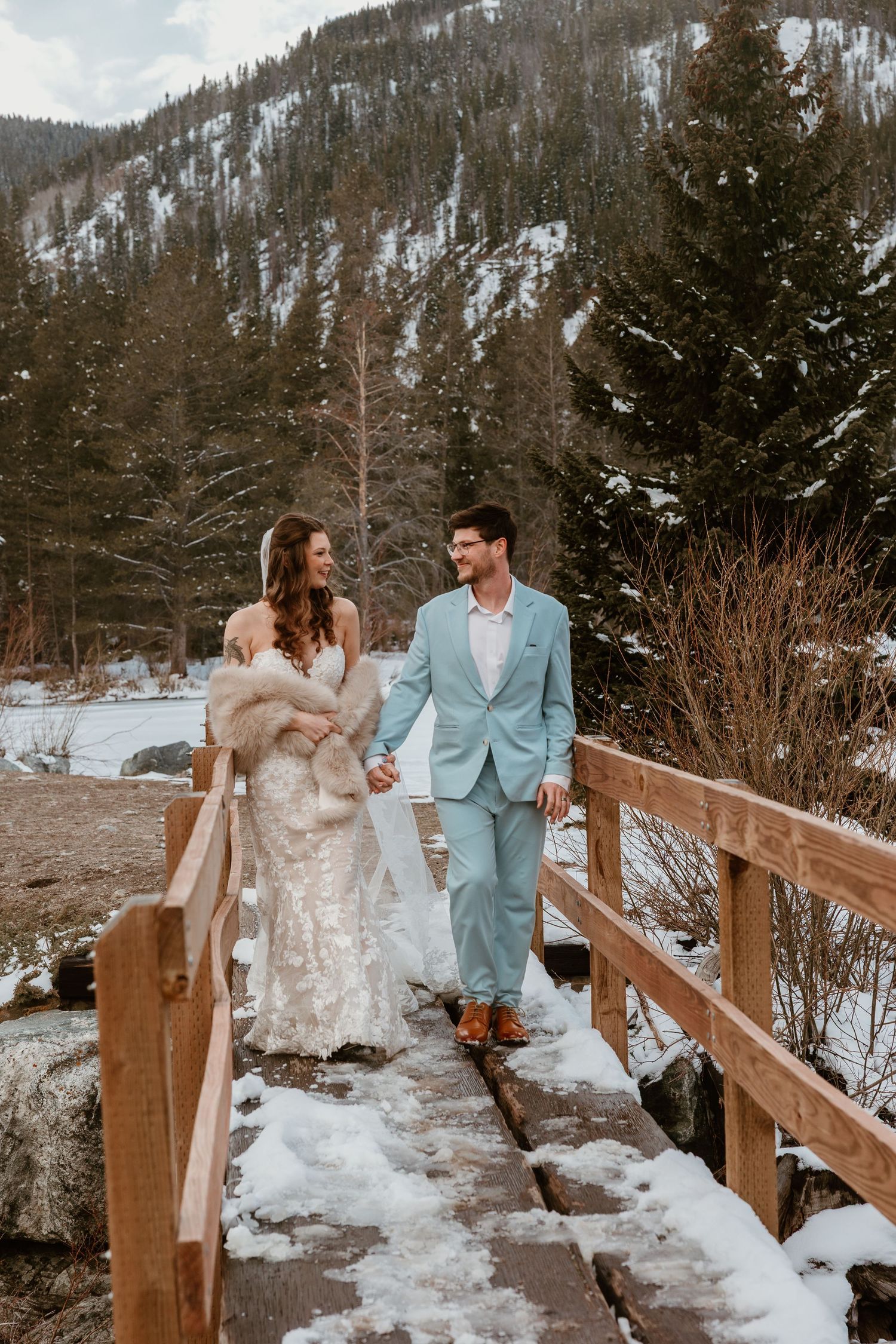 A winter wedding couple poses playfully on a snowy bridge with mountains in the background.