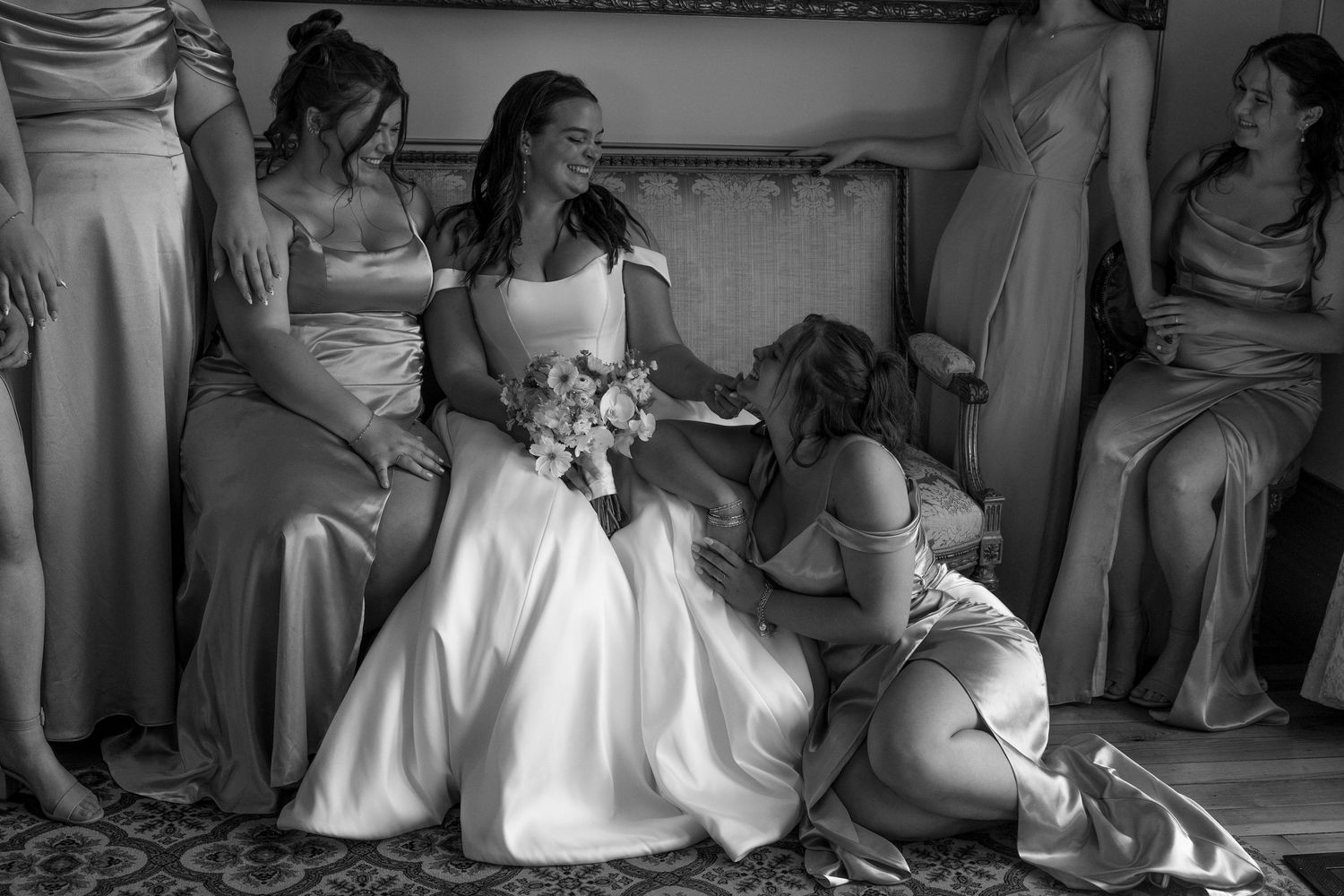 Wedding party members in white dresses and formal attire sit together on an elegant bench.