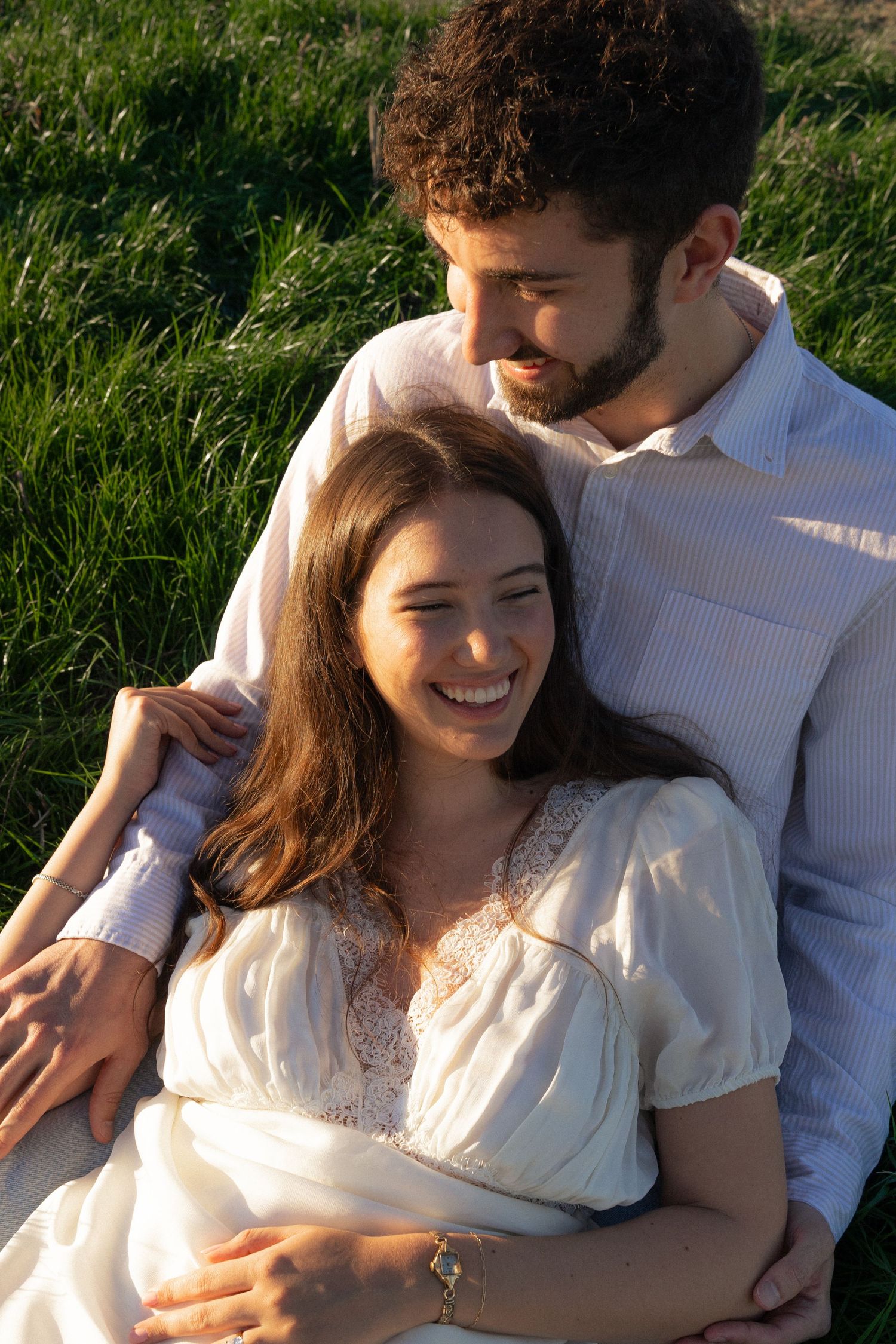 A couple in white clothing embraces while sitting in a sunlit grassy field during golden hour.