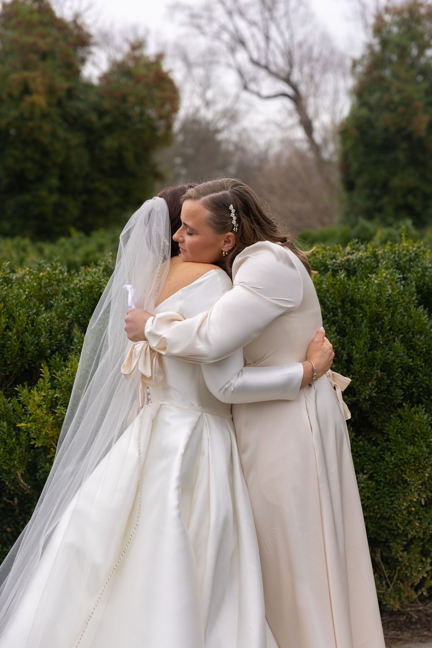 Two people in white wedding attire share an emotional embrace in front of green hedges on their special day.