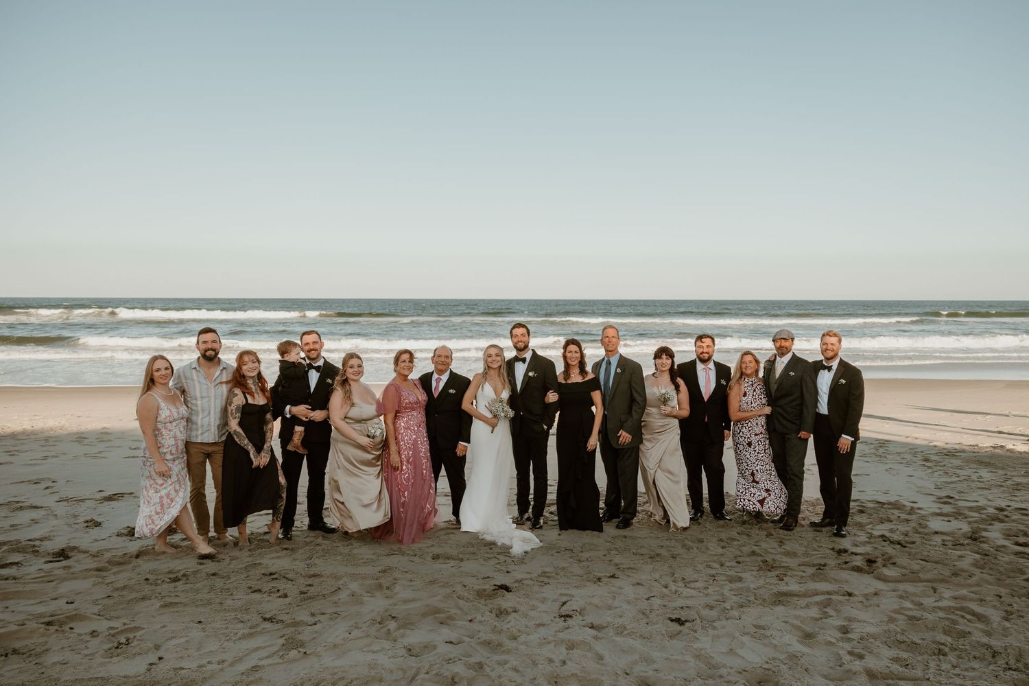 Wedding party group photo on beach with ocean waves and clear sky in background.