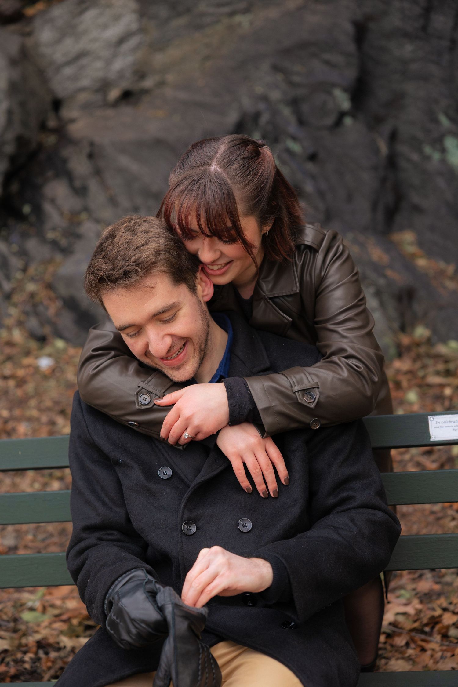 A couple shares a tender moment while sitting on a park bench surrounded by autumn leaves and rocky cliffs.