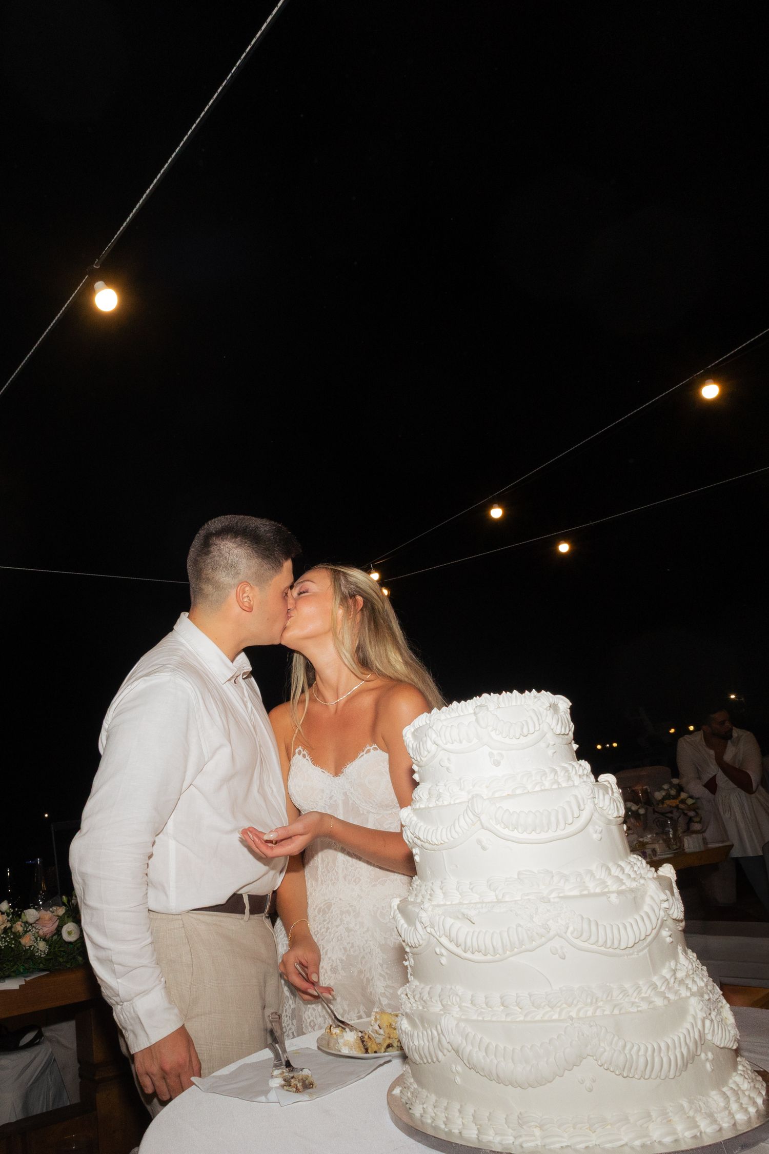 A couple shares a kiss beside their elegant white tiered wedding cake decorated with lace details under string lights at night.