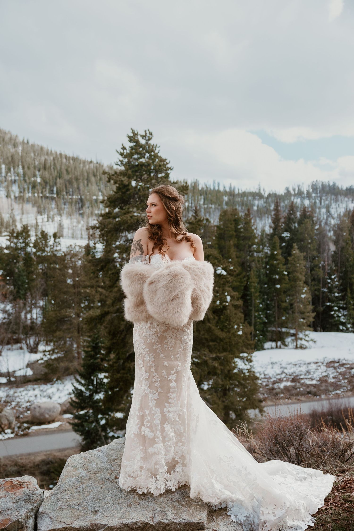 A bride in a lace dress and fur shawl poses on a rock with snowy mountains in the background.