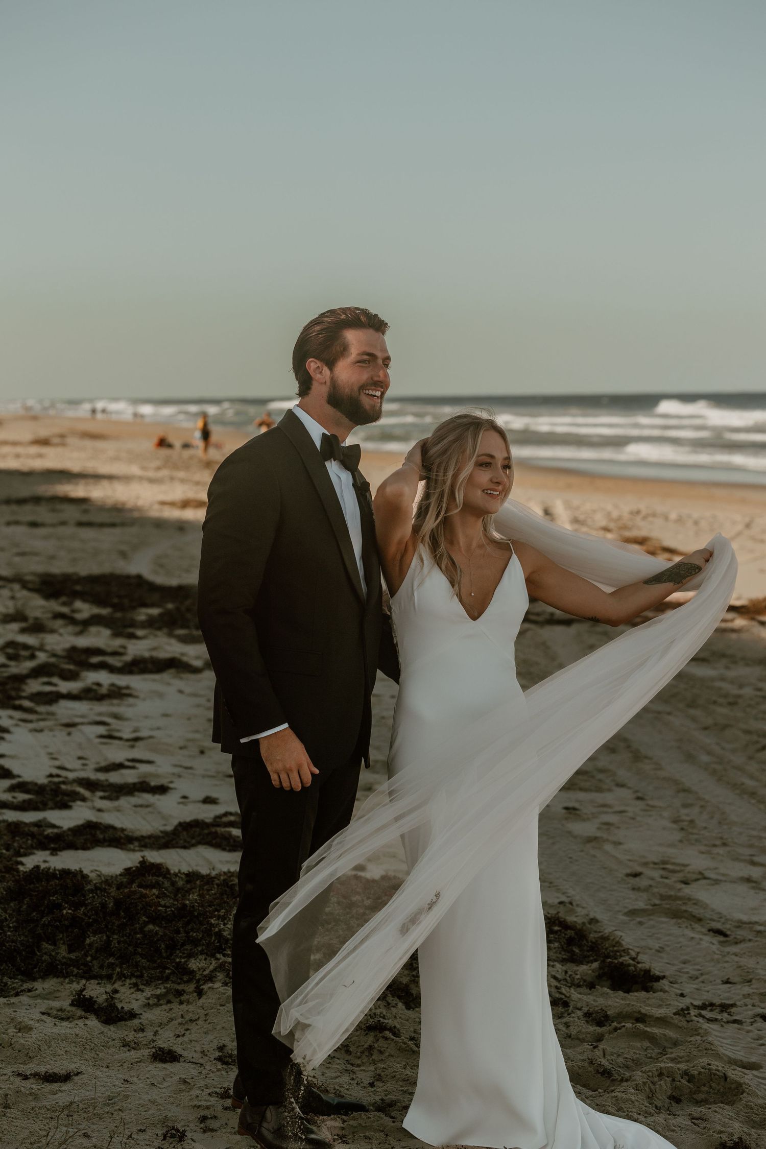 A couple poses on the beach at sunset with bride's veil flowing in the wind.