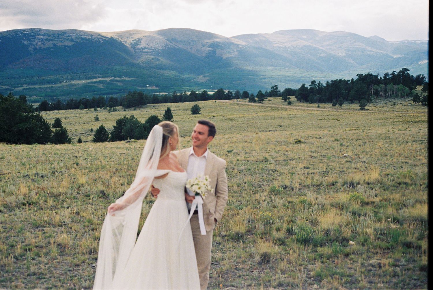Wedding couple embraces in a mountain field with dramatic mountain ranges in the distance.