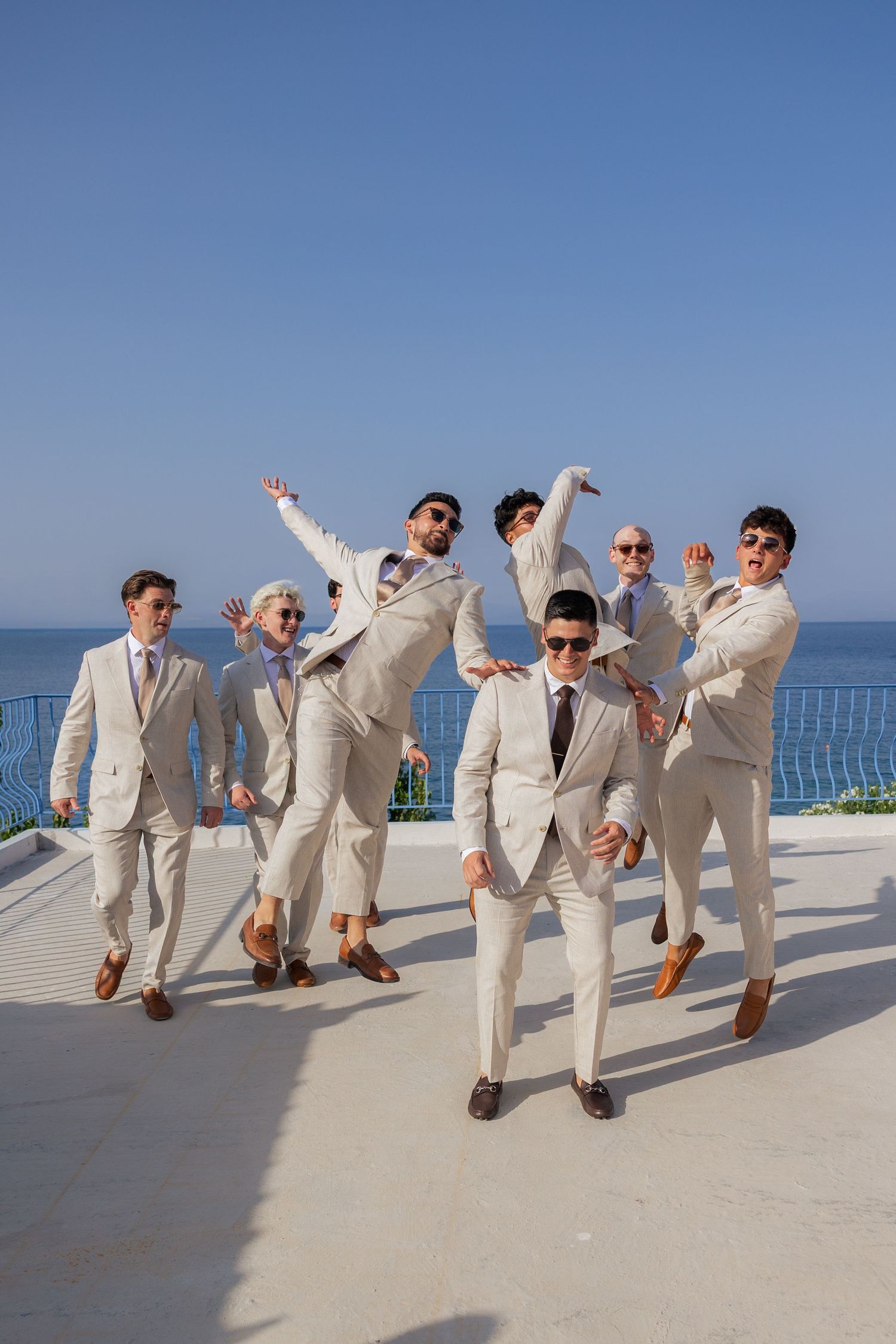 A group in light beige suits strike playful poses on a sunny terrace overlooking the ocean.