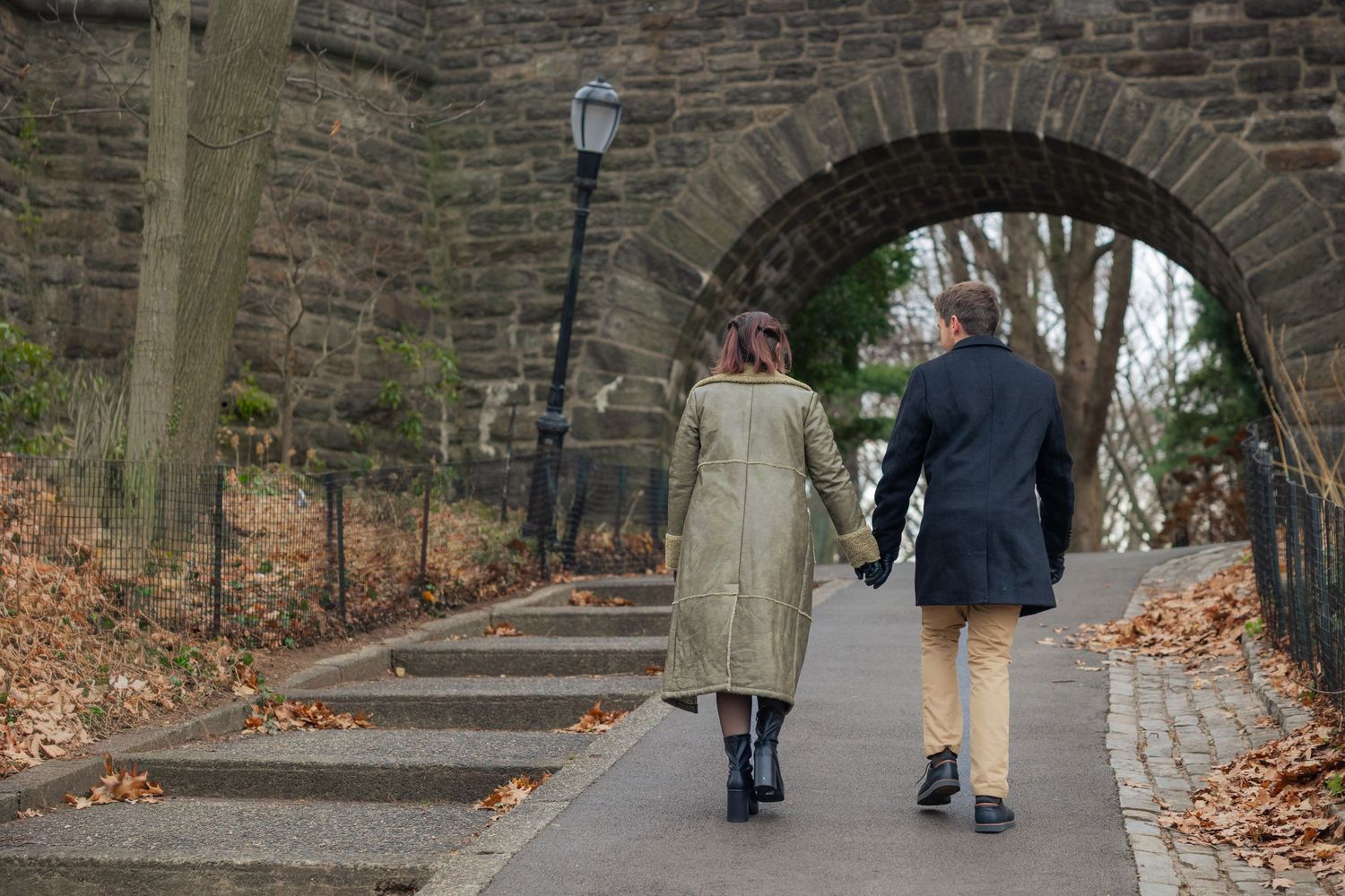 People walking through a stone archway tunnel in a park with fallen autumn leaves on the ground.