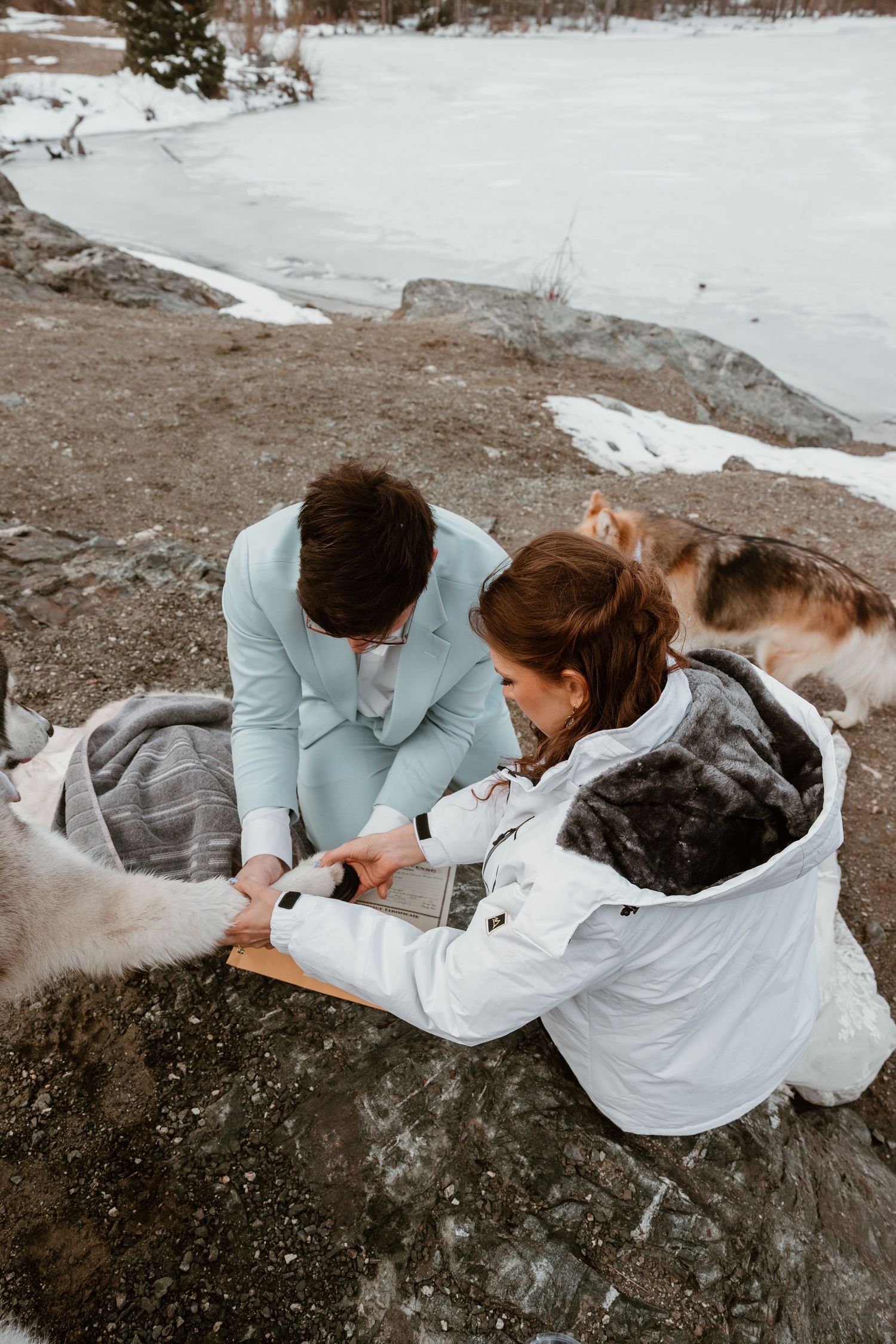 A couple in winter attire sits together on snowy ground during a proposal moment.