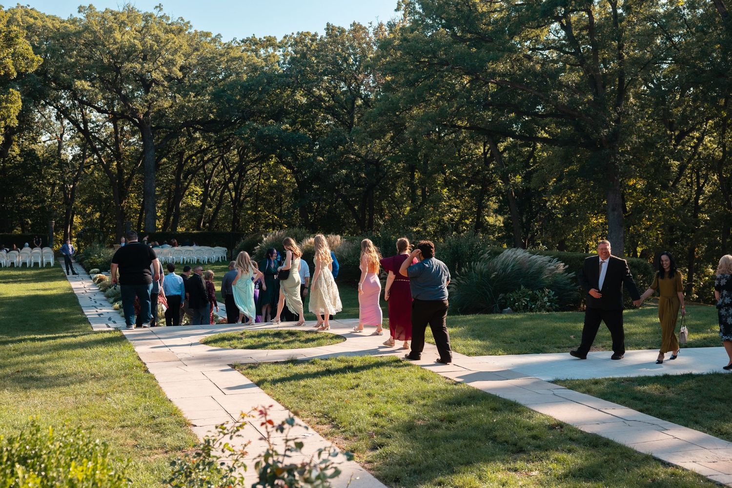 Wedding guests gather outdoors for a ceremony along a white walkway surrounded by lush green trees and grass.