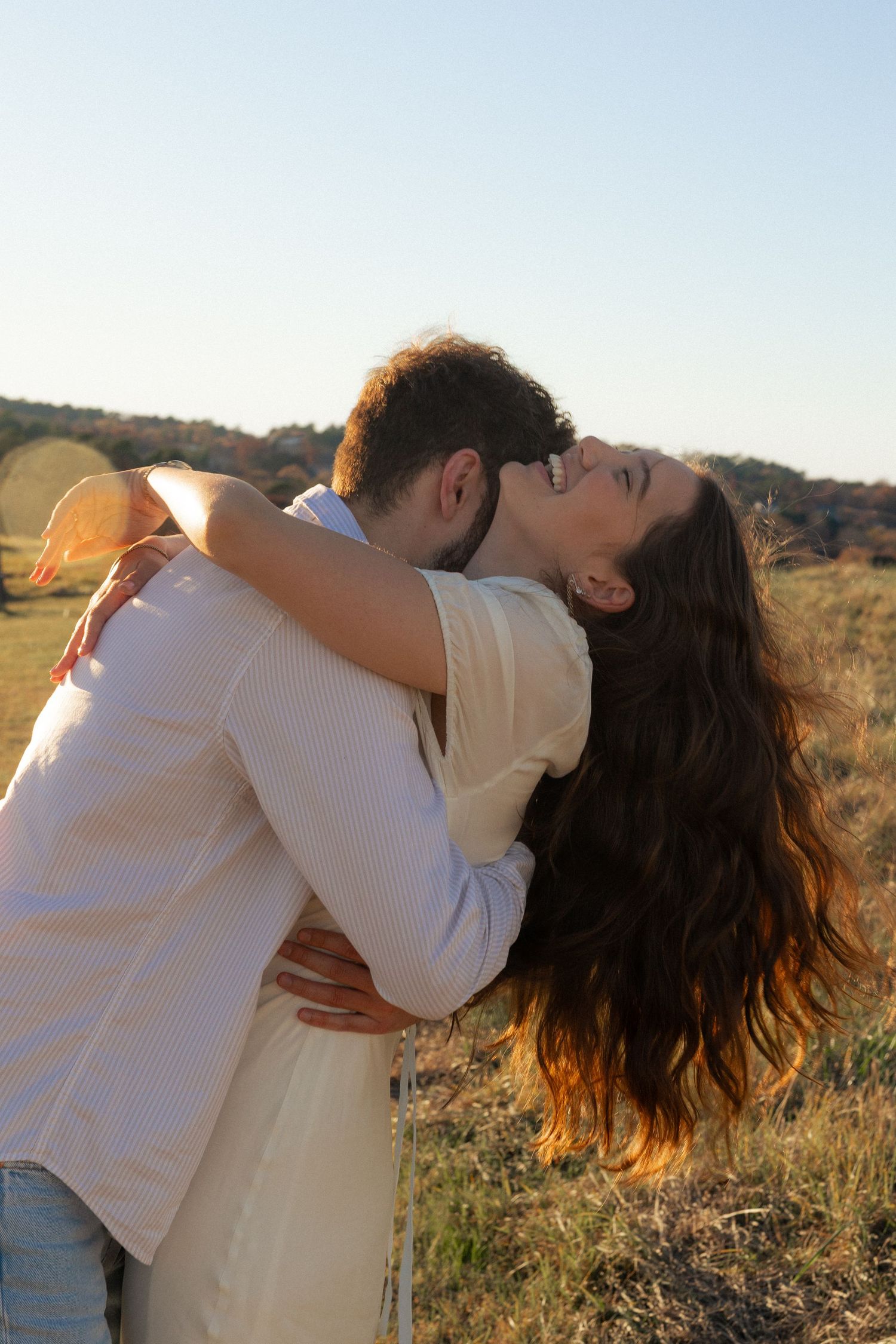 A romantic couple in light clothing share an intimate embrace against a sunset backdrop in a field.