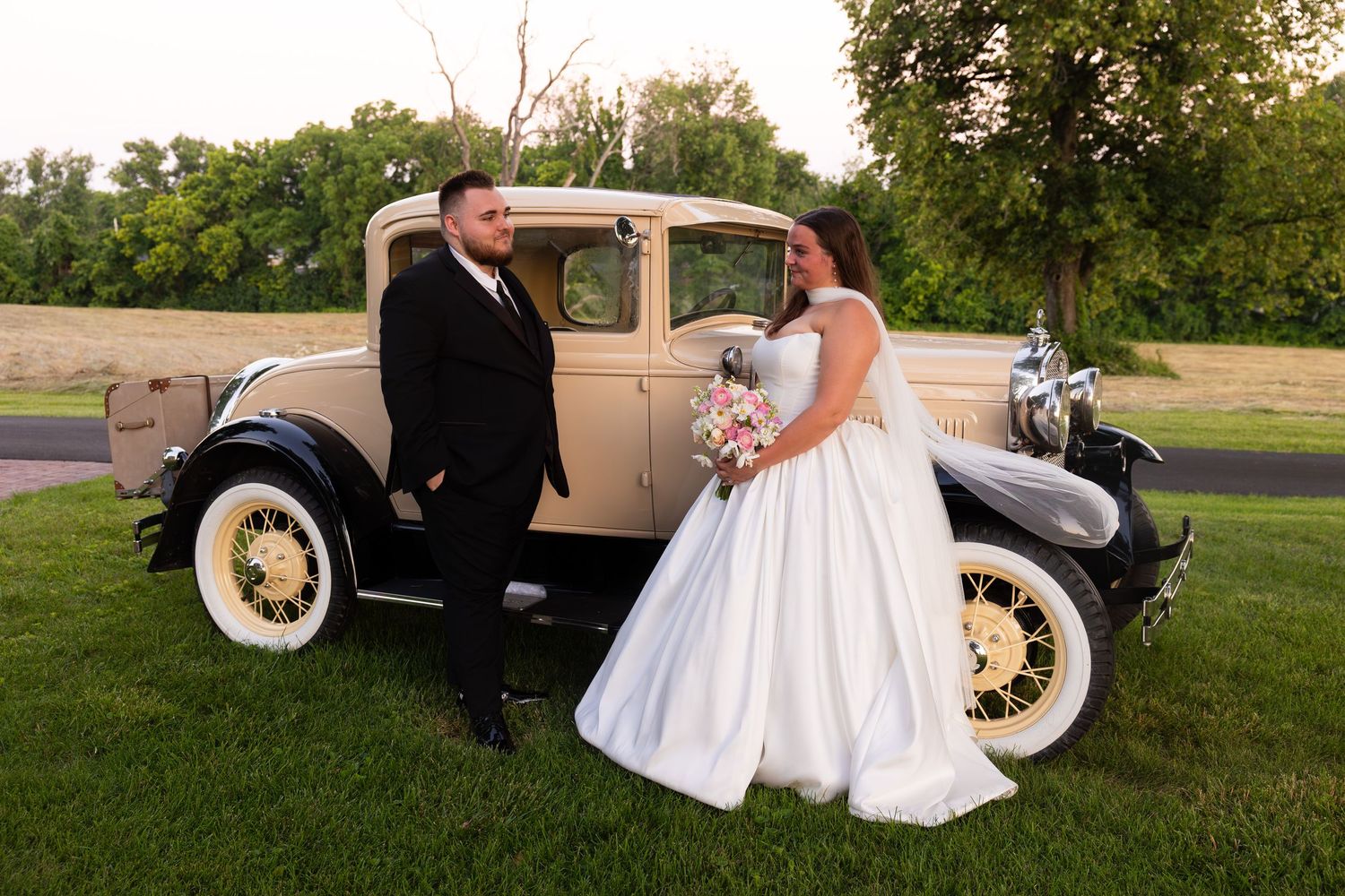 A couple poses next to a vintage cream-colored car on their wedding day in a grassy outdoor setting.