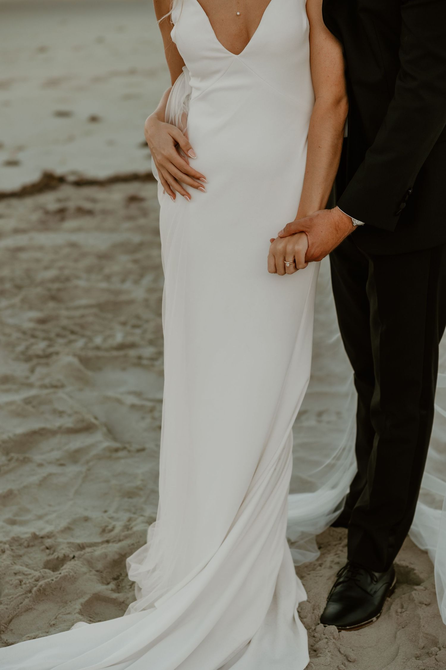 Close-up detail of an elegant white wedding dress and formal black suit on a beach.